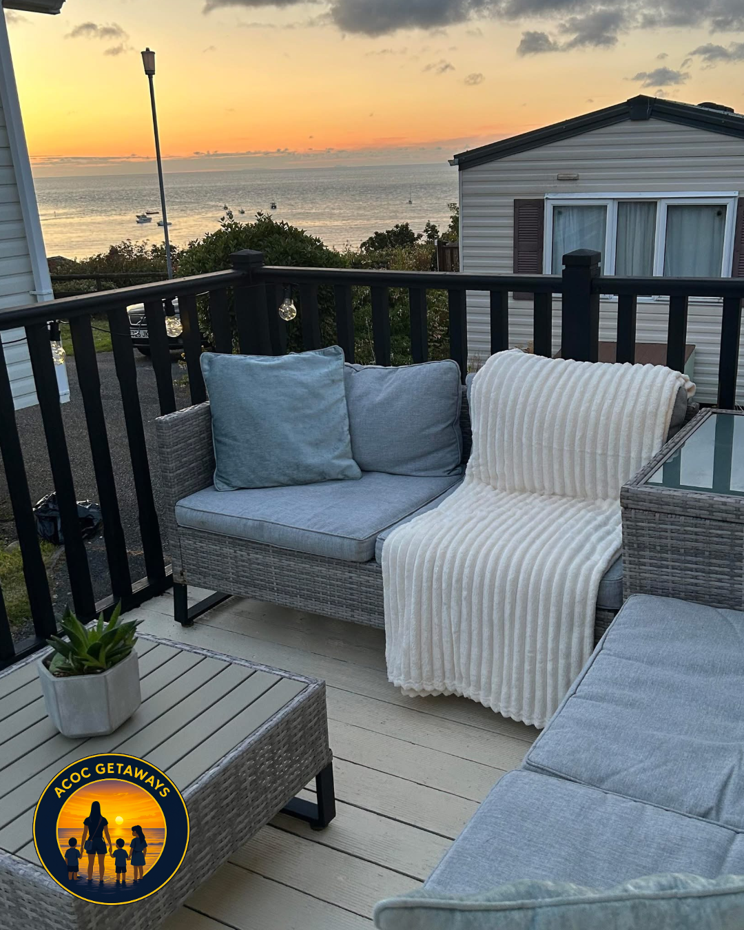 View of a balcony overlooking the ocean at sunset with a wicker sofa, gray cushions, a cream blanket, a small table with a potted plant, and houses in the background.