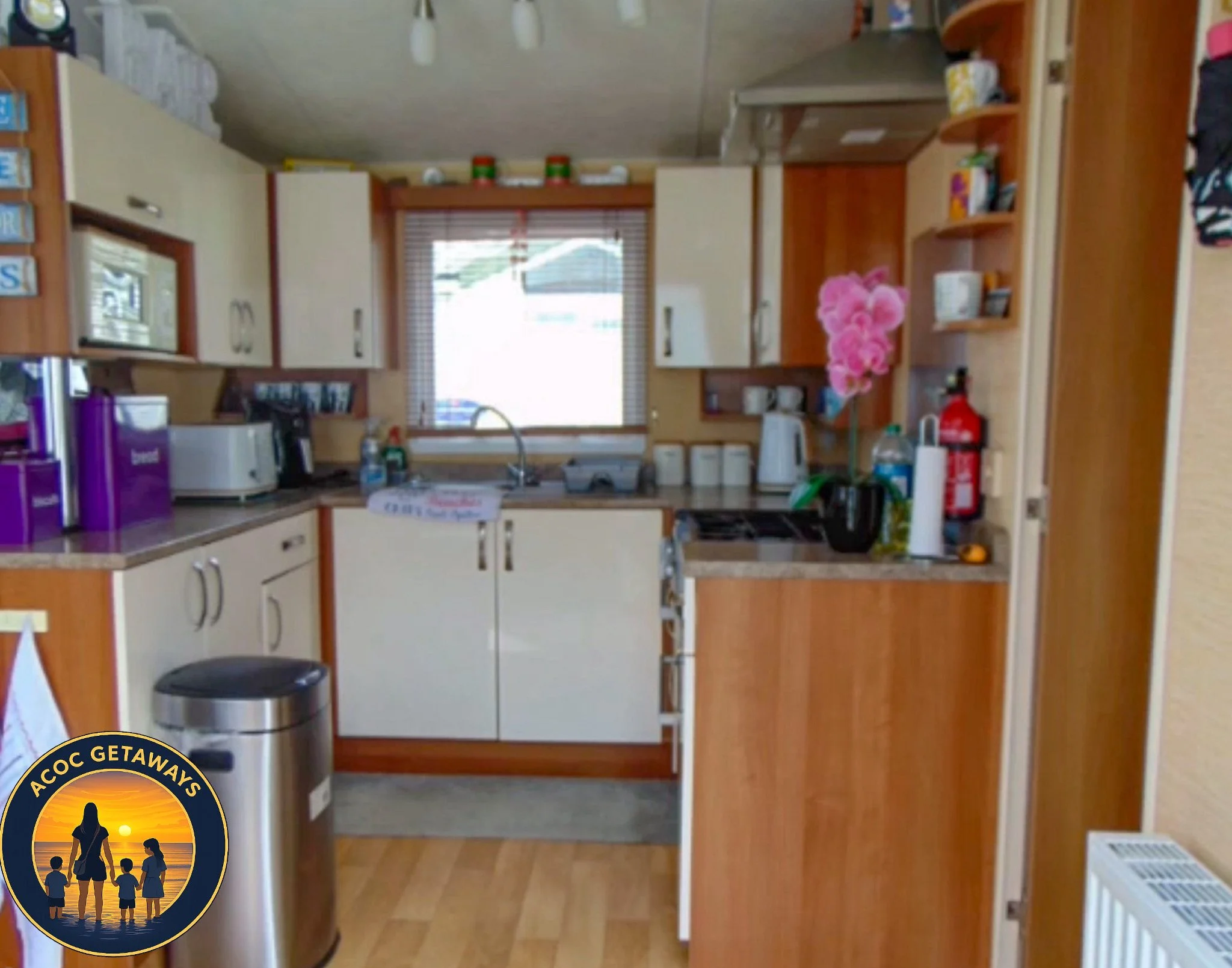 View of a compact kitchen with wooden cabinets, a window above the sink, and various appliances including a microwave, toaster, and coffee maker. There are items on the counter, a fire extinguisher on the wall, and pink flowers in a vase. A laundry h