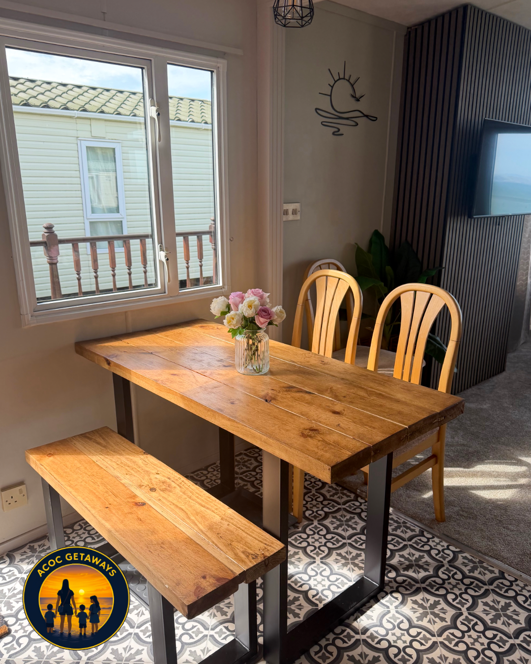 A wooden dining table with three chairs and a bench, decorated with a vase of pink and white flowers. The table is next to a window with a view of neighboring houses. The room has patterned tile flooring and a wall with a sun and moon wall decoration