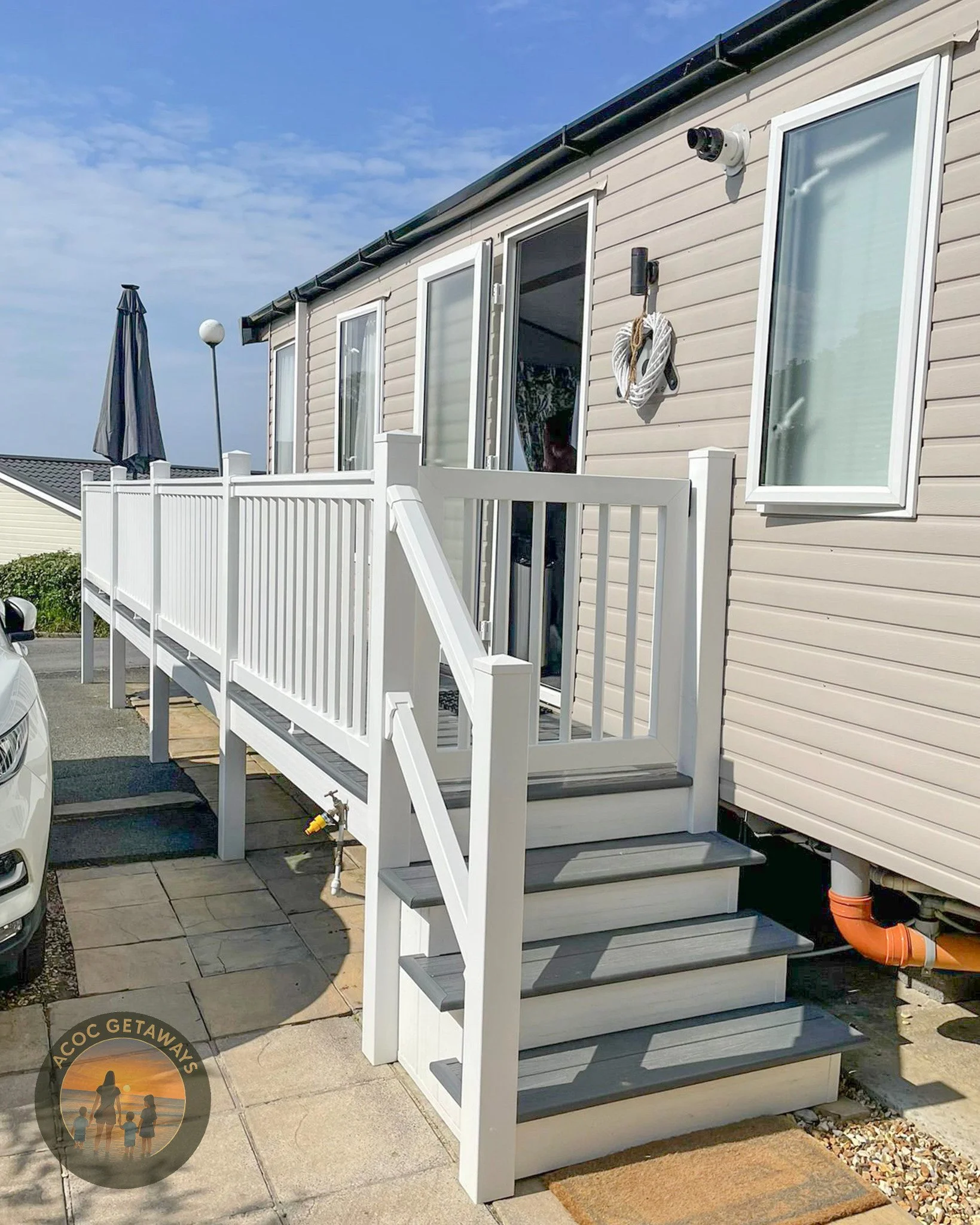 Exterior view of a house with a white staircase leading to a sliding glass door on the porch. The house has beige siding, and there is a CCTV camera mounted above the door. An outdoor umbrella is visible in the background, and a streetlamp is nearby.
