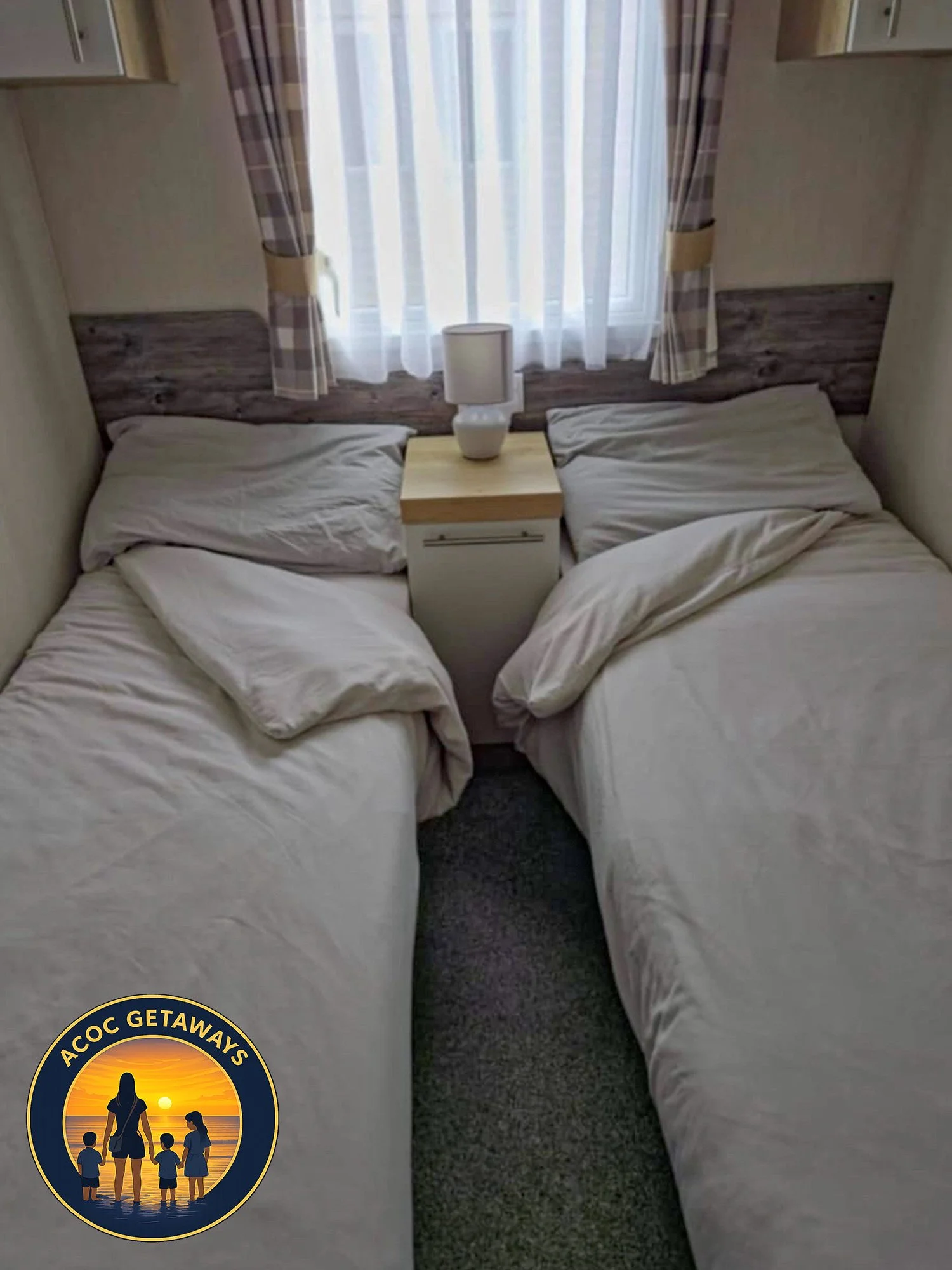 Two unmade twin beds with white bedding, separated by a nightstand with a lamp, under a window with sheer curtains and plaid drapes.