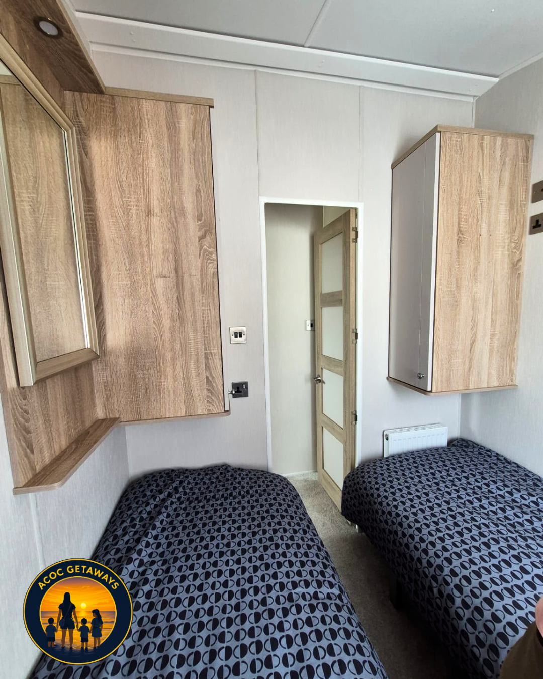 A small bedroom with two single beds with black geometric patterned bedding, wooden wall-mounted cabinets, a small mirror, a door, and a radiator.