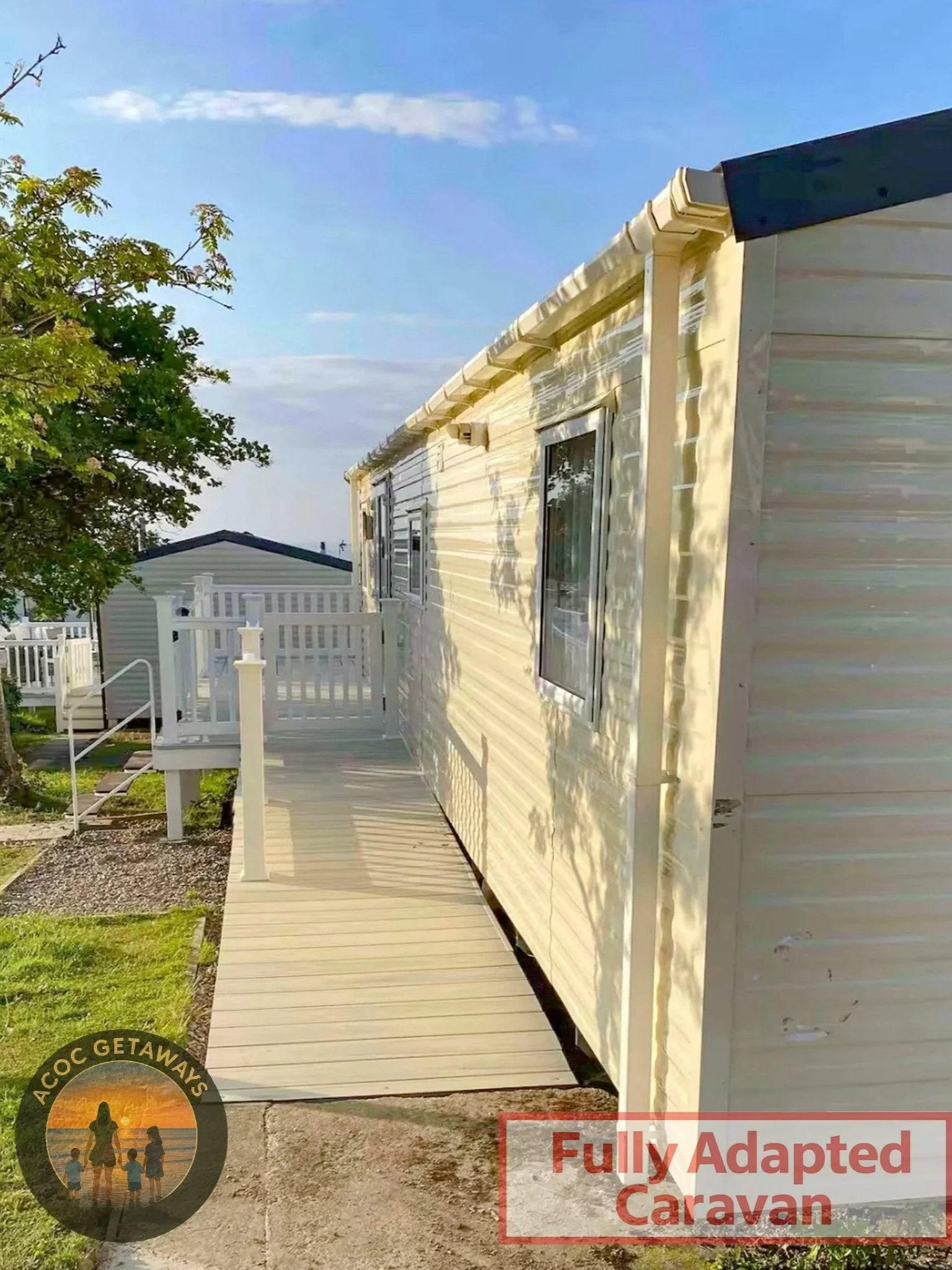 Exterior side of a fully adapted caravan with a small wooden porch and railing, surrounded by grass and trees, under a blue sky with some clouds.
