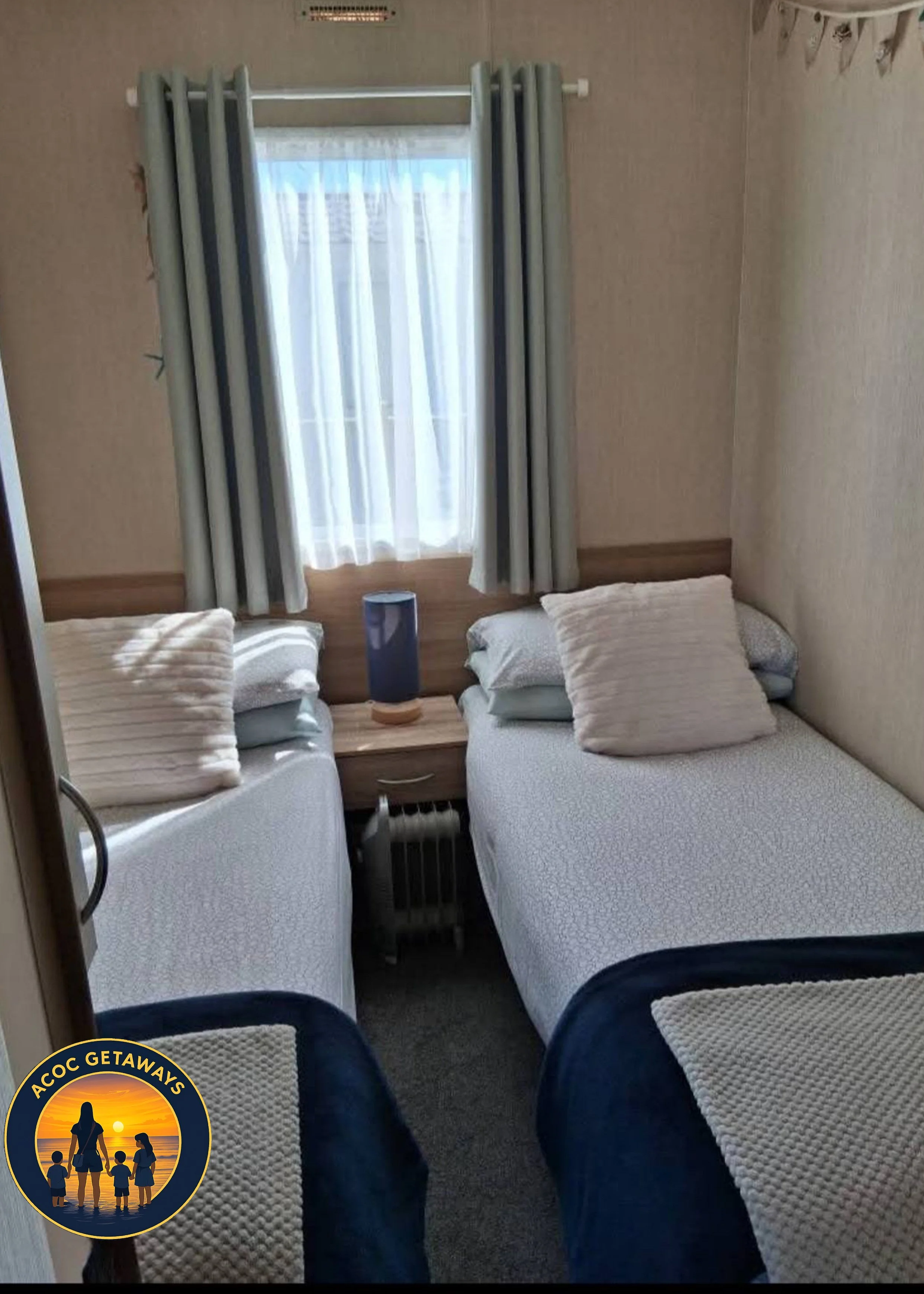 A small hotel room with two twin beds, pillows, a nightstand between them, a lamp, and a window with curtains. There is a radiator beneath the window and a logo in the bottom left corner with a sunset and silhouettes of a family.