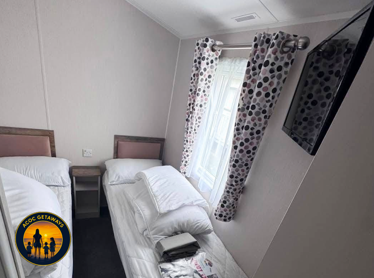 A bedroom with two single beds, each with white bedding and pillows, a small wooden nightstand positioned between them, a window with patterned curtains, and a flat-screen TV mounted on the wall. There is also a stack of items, possibly magazines or 