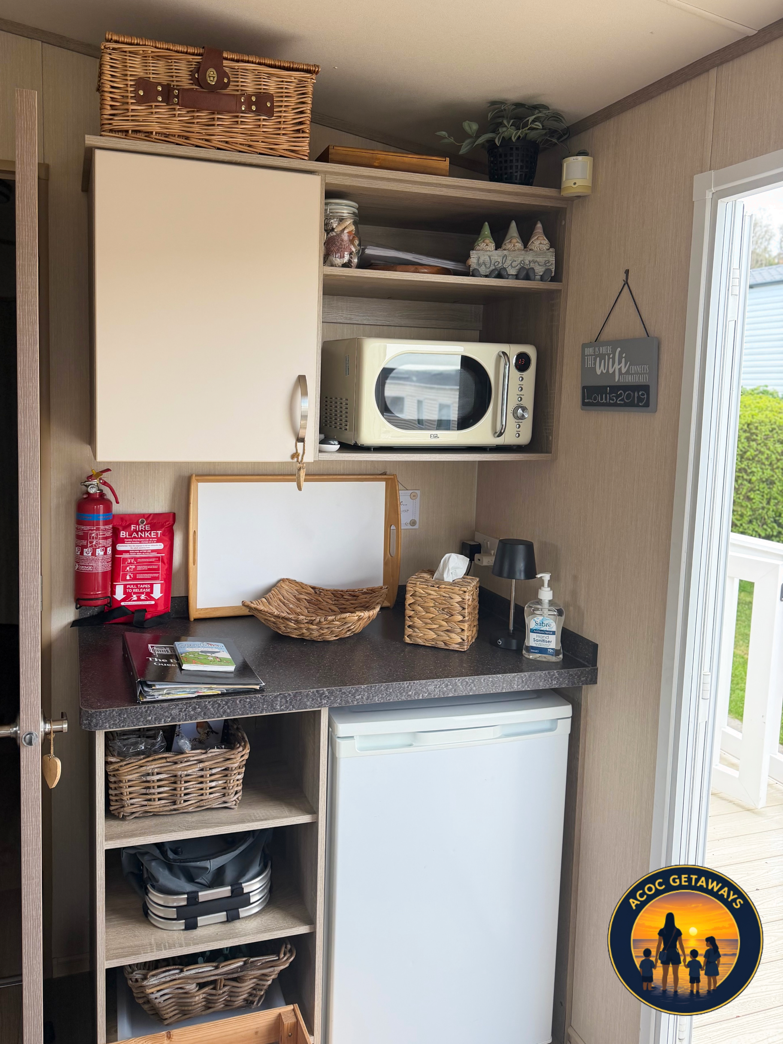 A small kitchen or pantry corner with a wooden shelf holding a woven basket, some decorative gnomes, and potted plant. Below the shelf is a microwave oven and a counter with wicker baskets, a tissue box, and a hand sanitizer. To the side, there is a 
