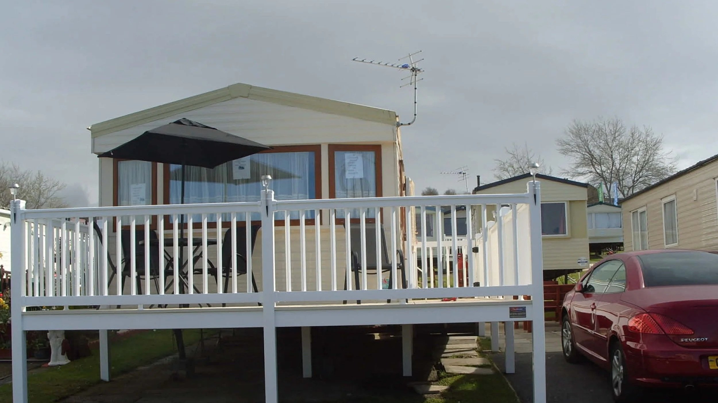 A mobile home with a deck, black umbrella, and outdoor furniture. A red car is parked nearby. Several other mobile homes are visible in the background.