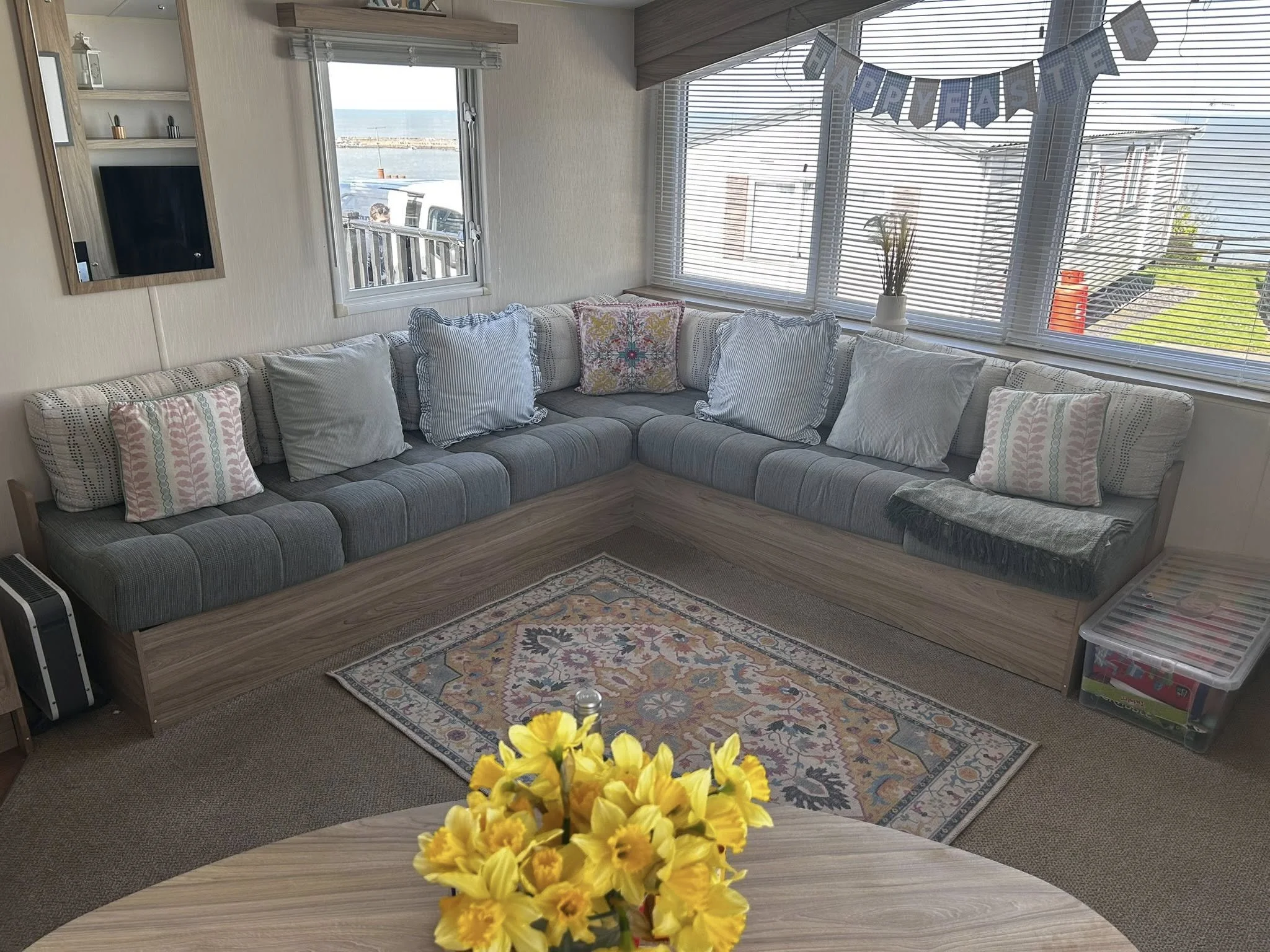 A cozy living room with a sectional sofa decorated with assorted pastel pillows, a colorful area rug, large windows with blinds, and ocean view through the windows. A 