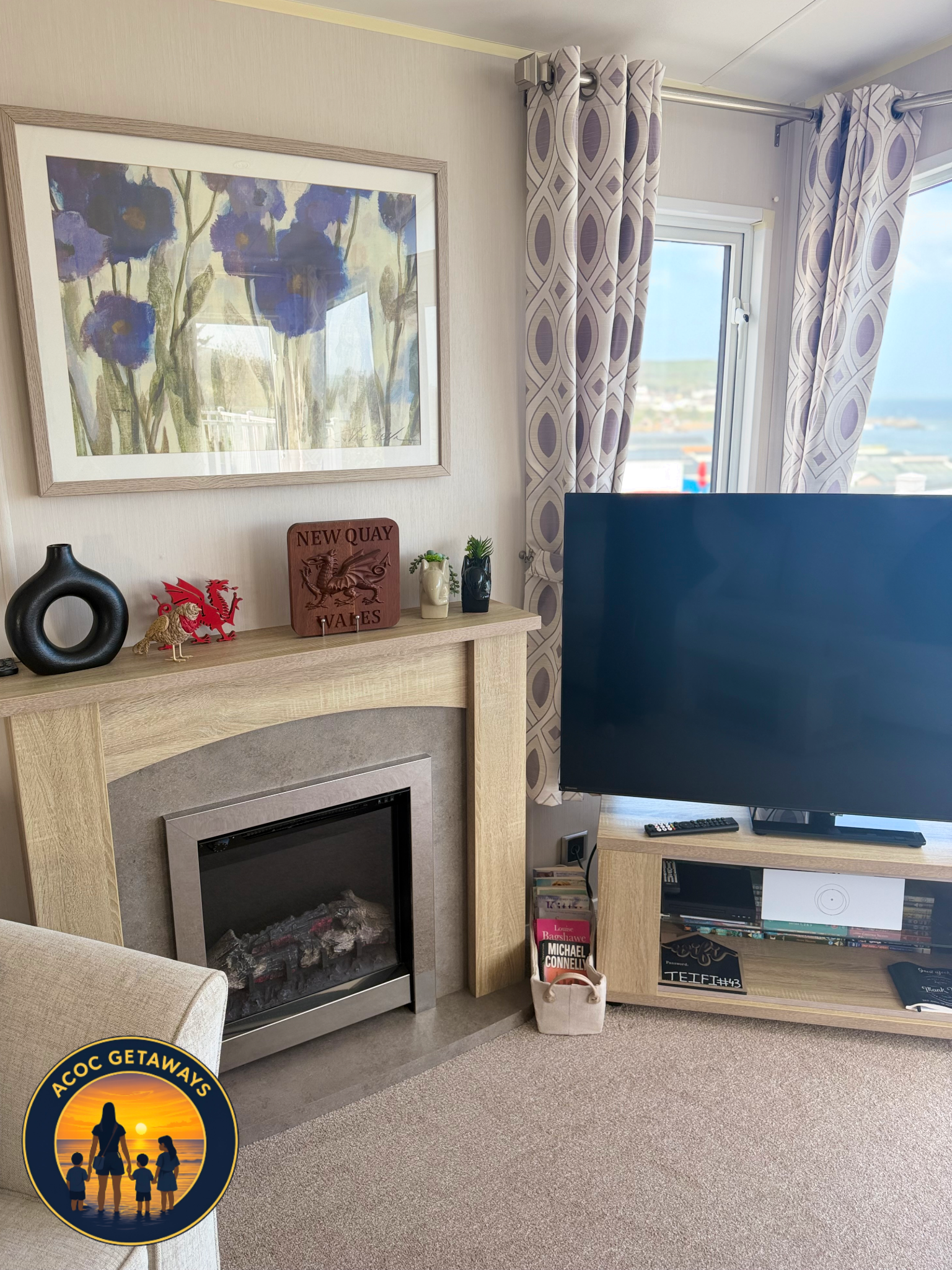 A living room with a beige fireplace, colorful decorations, and a flat-screen TV. A window with patterned curtains shows a view of the outdoors, including water and distant land. Items on the mantel include a black vase, red dragon, wooden sign readi