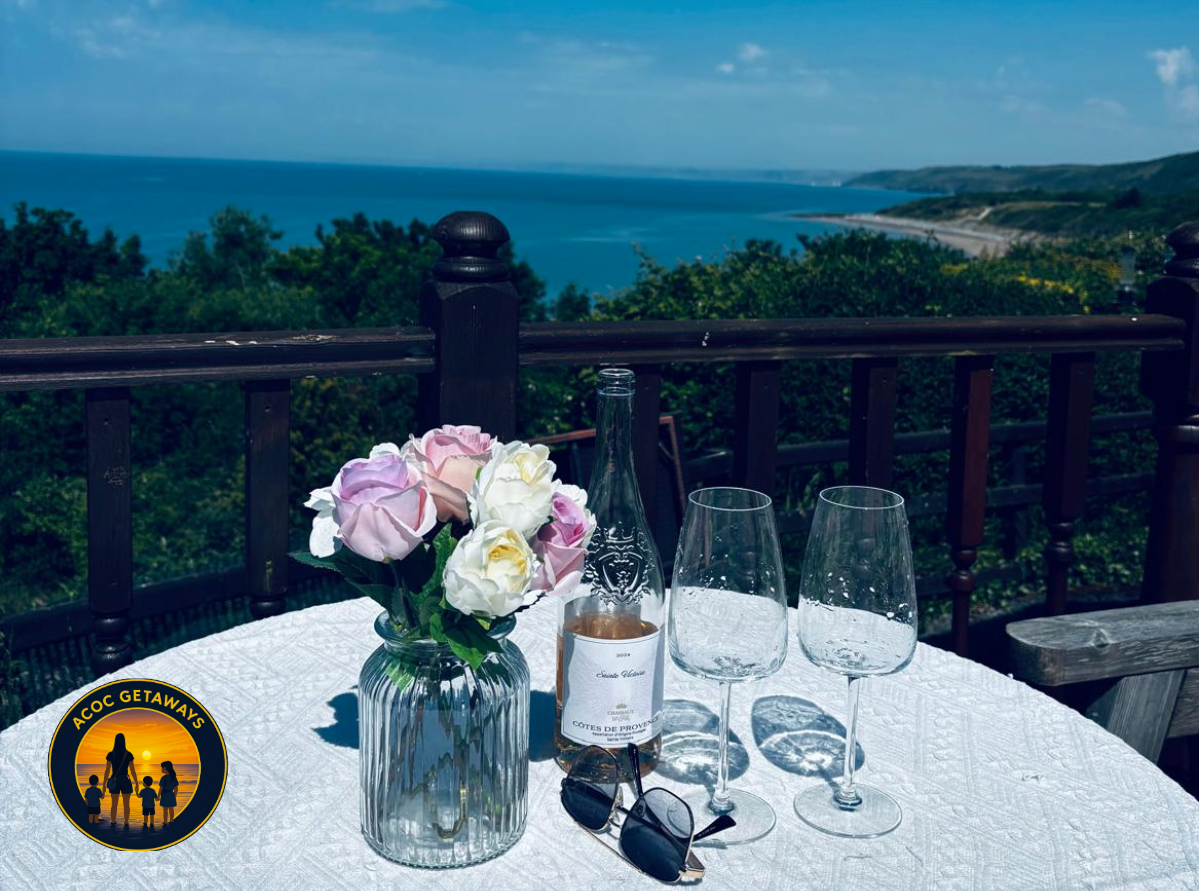 A table with a white tablecloth, a vase of pink and white flowers, a wine bottle, two empty wine glasses, and a pair of sunglasses, with a scenic view of the ocean and greenery in the background.