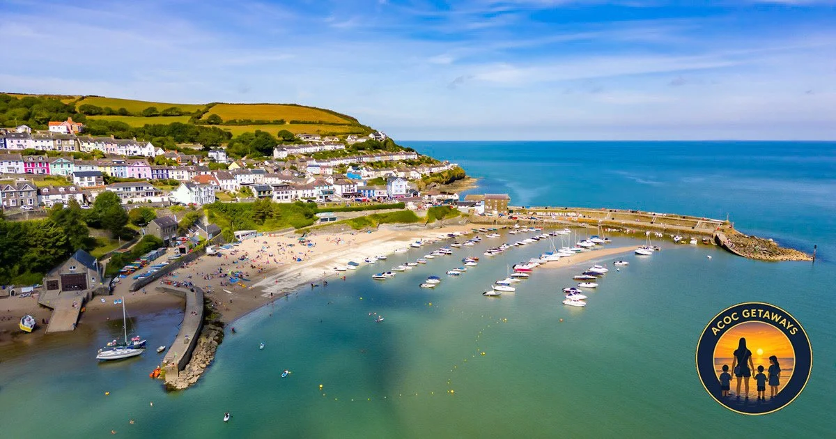 Aerial view of a coastal town with a sandy beach filled with people, colorful houses on the hillside, boats in the harbor, and the ocean extending to the horizon. An inset logo with a sunset scene and the text "Acoce Getaways" featuring a family silhouette.