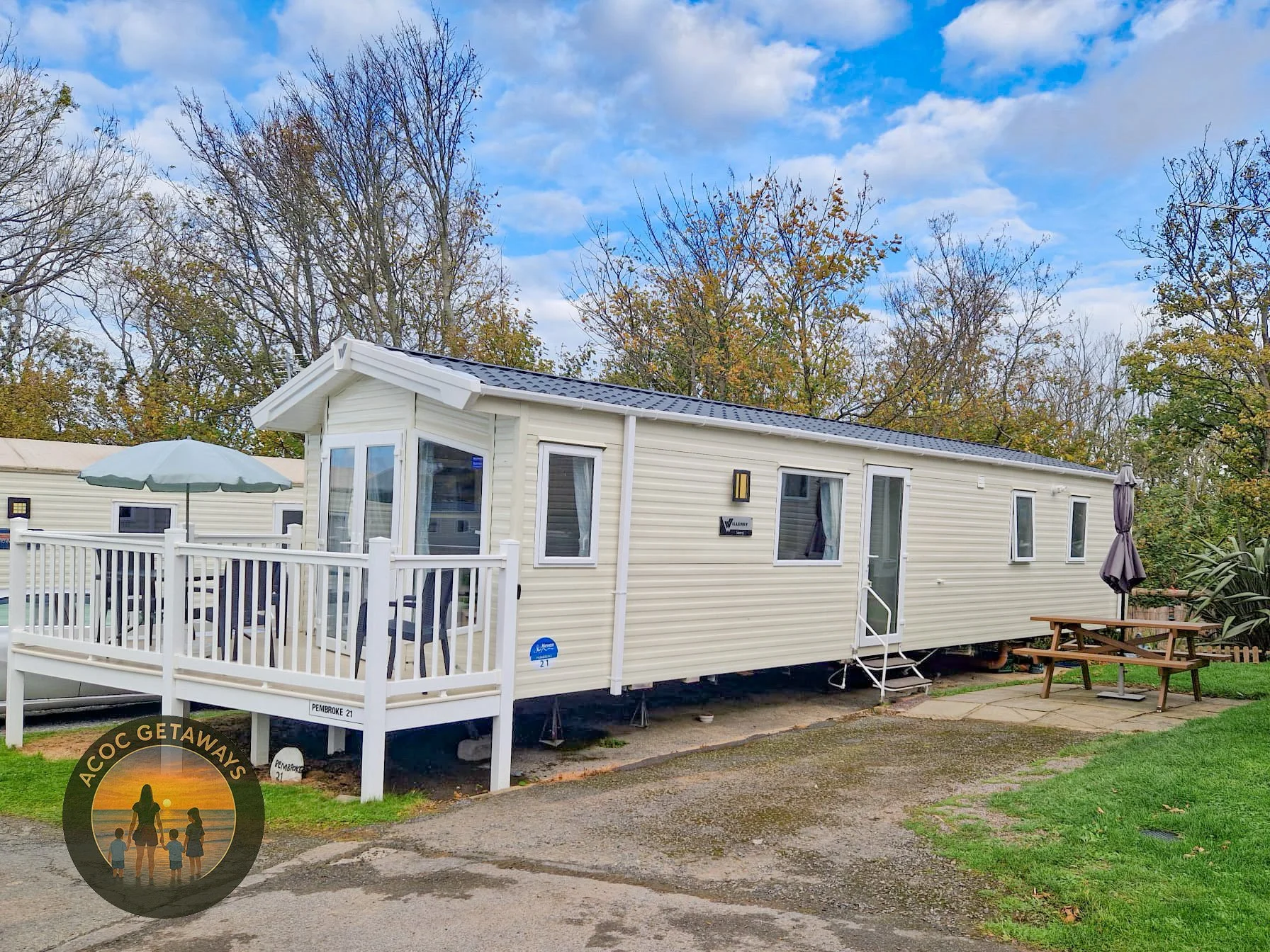 A modern, white mobile home with a small porch and outdoor seating area, surrounded by trees with autumn foliage, and a partly cloudy sky overhead.