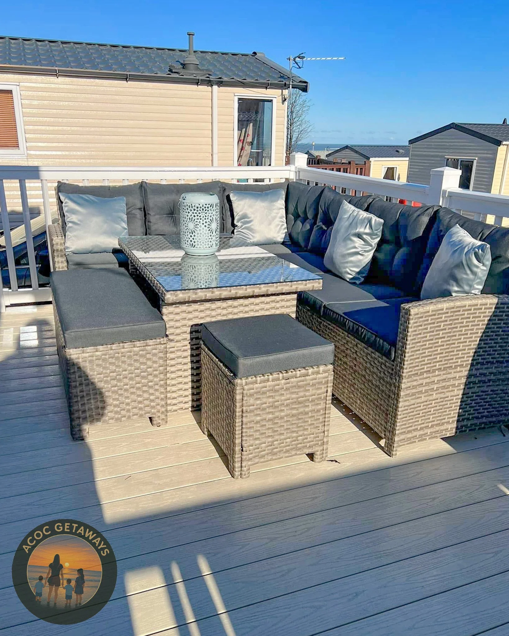 Outdoor patio with a wicker sectional sofa with dark cushions and silver pillows, a wicker ottoman, and a woven lantern on a glass-topped wicker coffee table, all on a wooden deck with a white railing, under a clear blue sky.