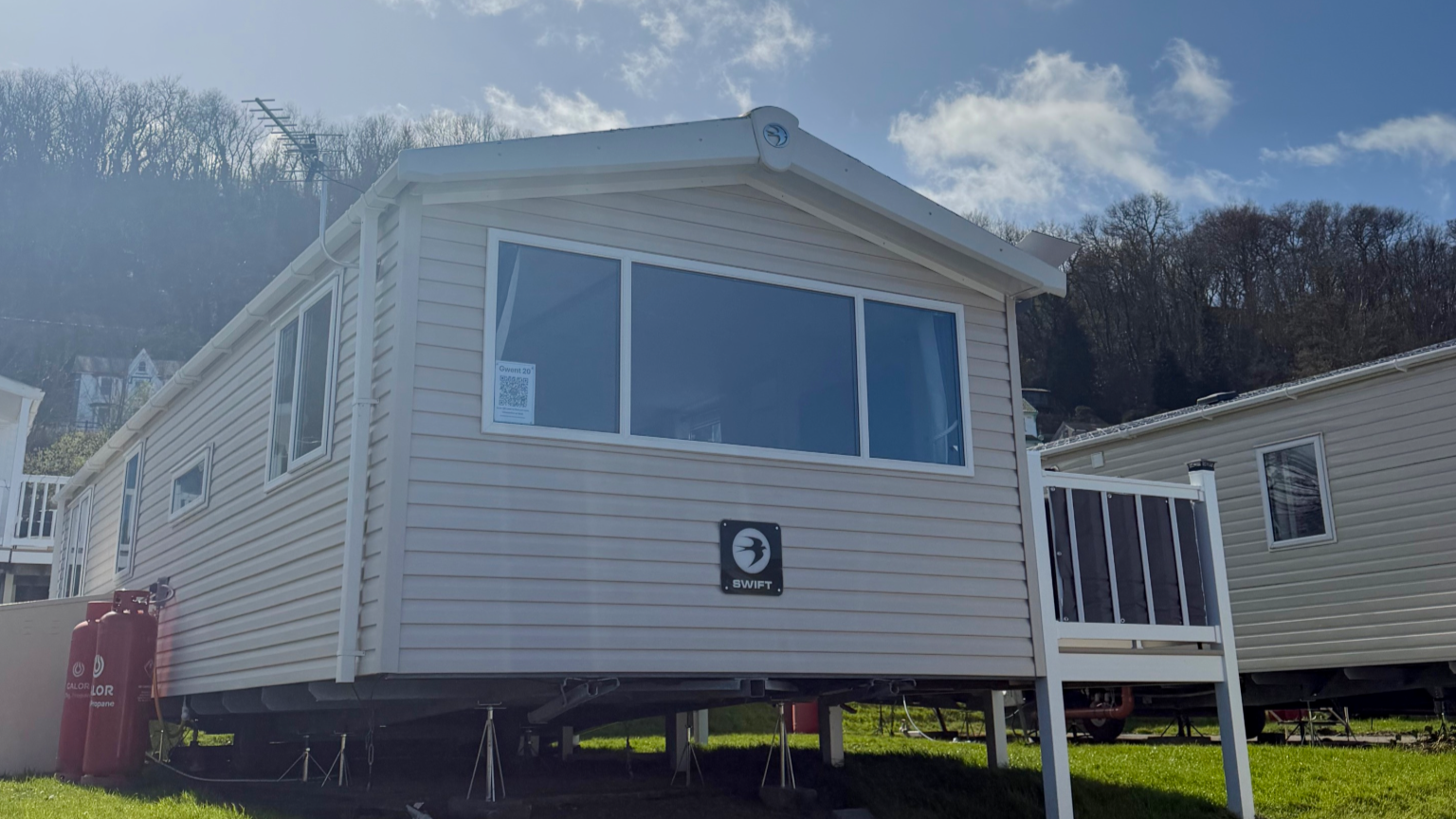 A white modular home with a large front window and a small side window, built on stilts on a grassy area. The house has a logo with the word 'SWIFT' on the front. There are propane tanks to the left of the house and another similar house to the right. Trees and a blue sky with scattered clouds are in the background.
