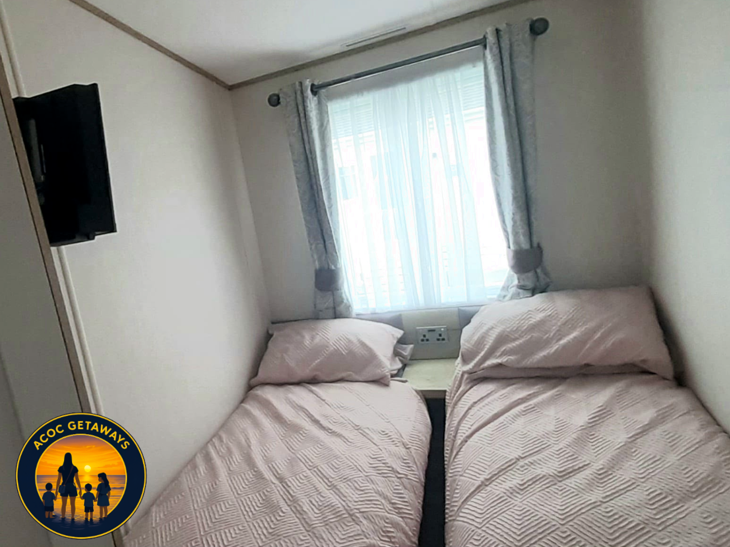 A small bedroom with two single beds covered in light pink bedspreads, a window with white curtains and blue floral drapes, and a wall-mounted TV opposite the beds.