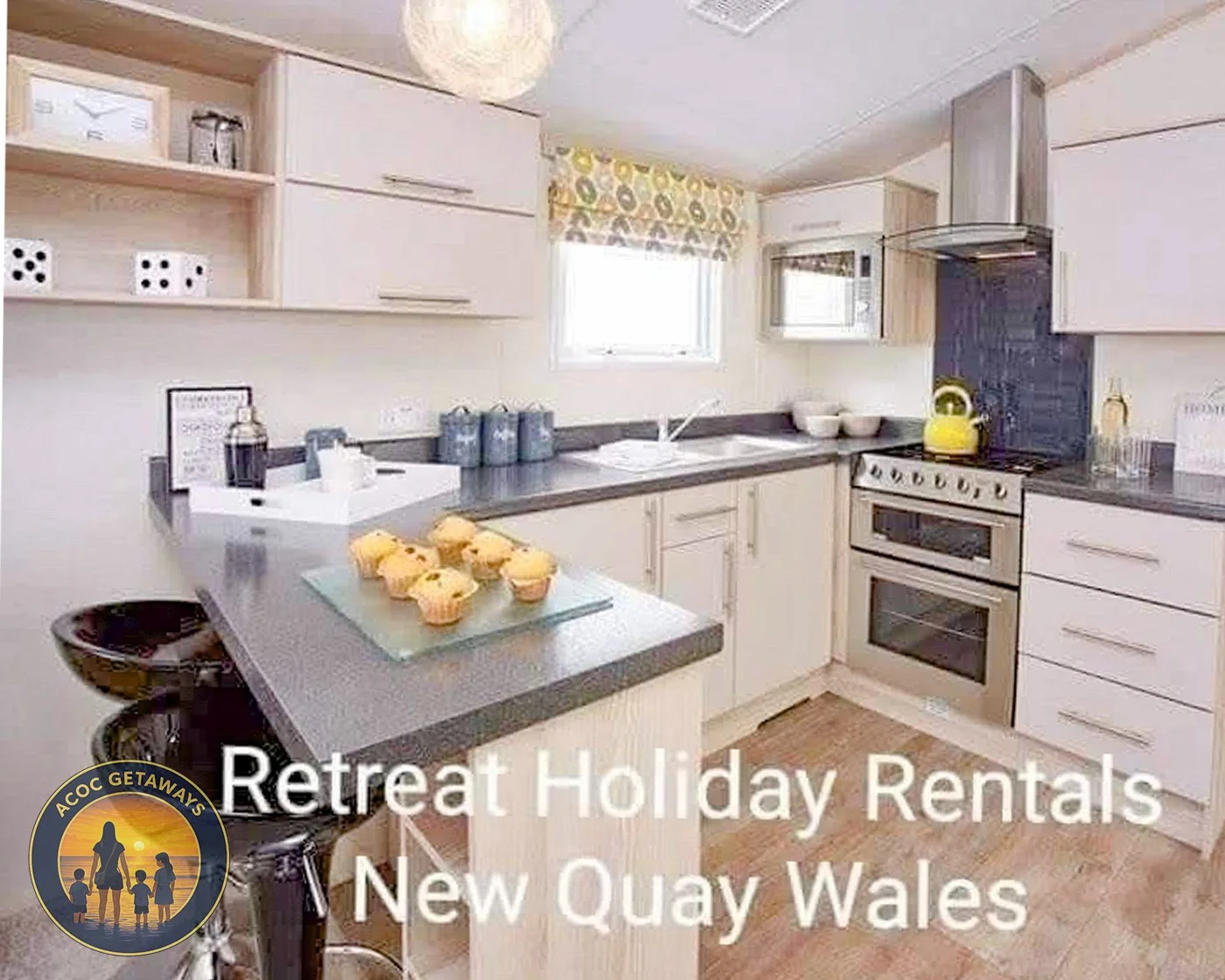 Cozy kitchen with white cabinets, gray countertops, yellow teapot, microwave, oven, window with patterned curtain, and cupcakes on the counter. Text overlay reads 'Retreat Holiday Rentals New Quay Wales' with a logo of a family at sunset.