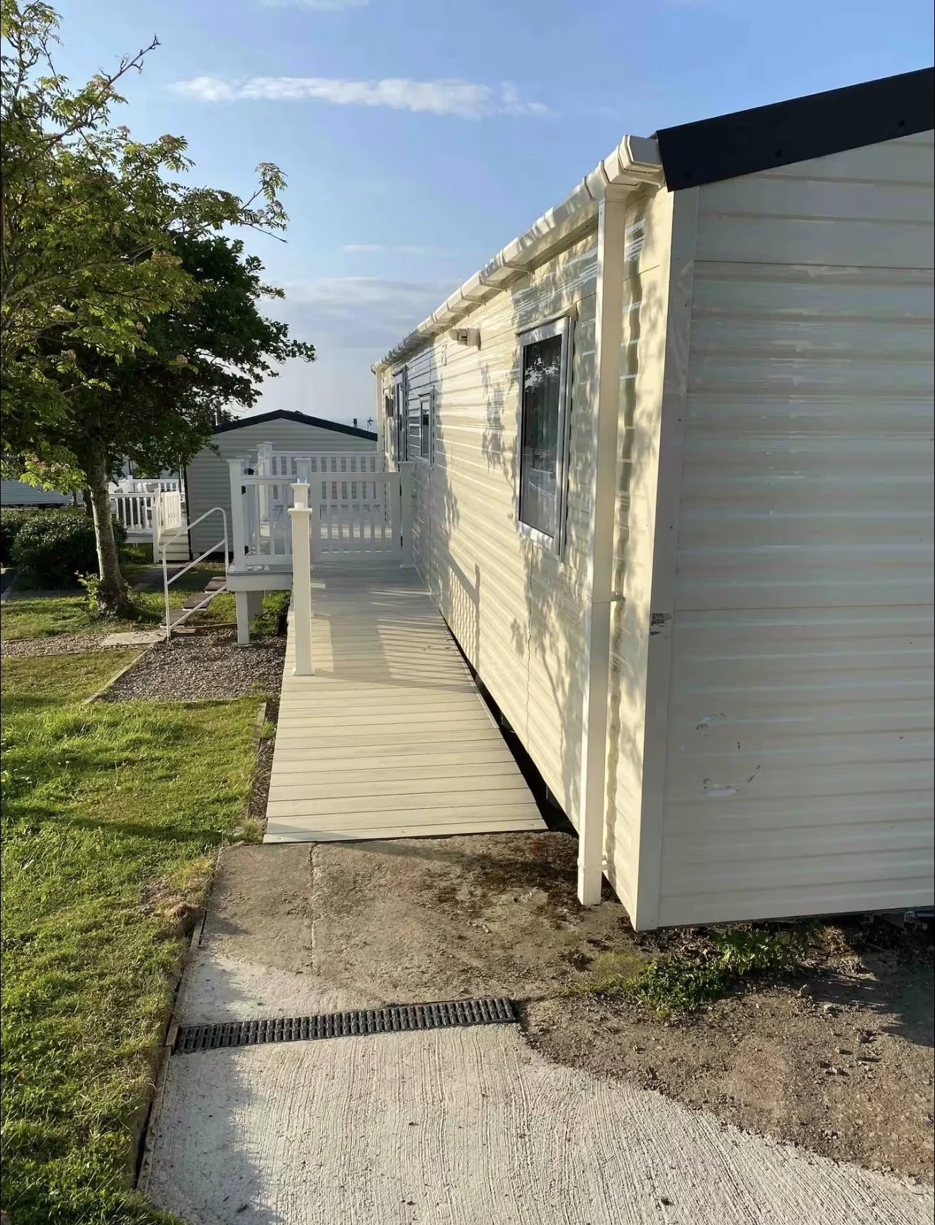 A mobile home with a white exterior, a small porch with white railings, and a wooden ramp leading to the entrance. There is a tree with green leaves on the left side and a grassy yard with a concrete walkway. The sky is blue with some clouds.