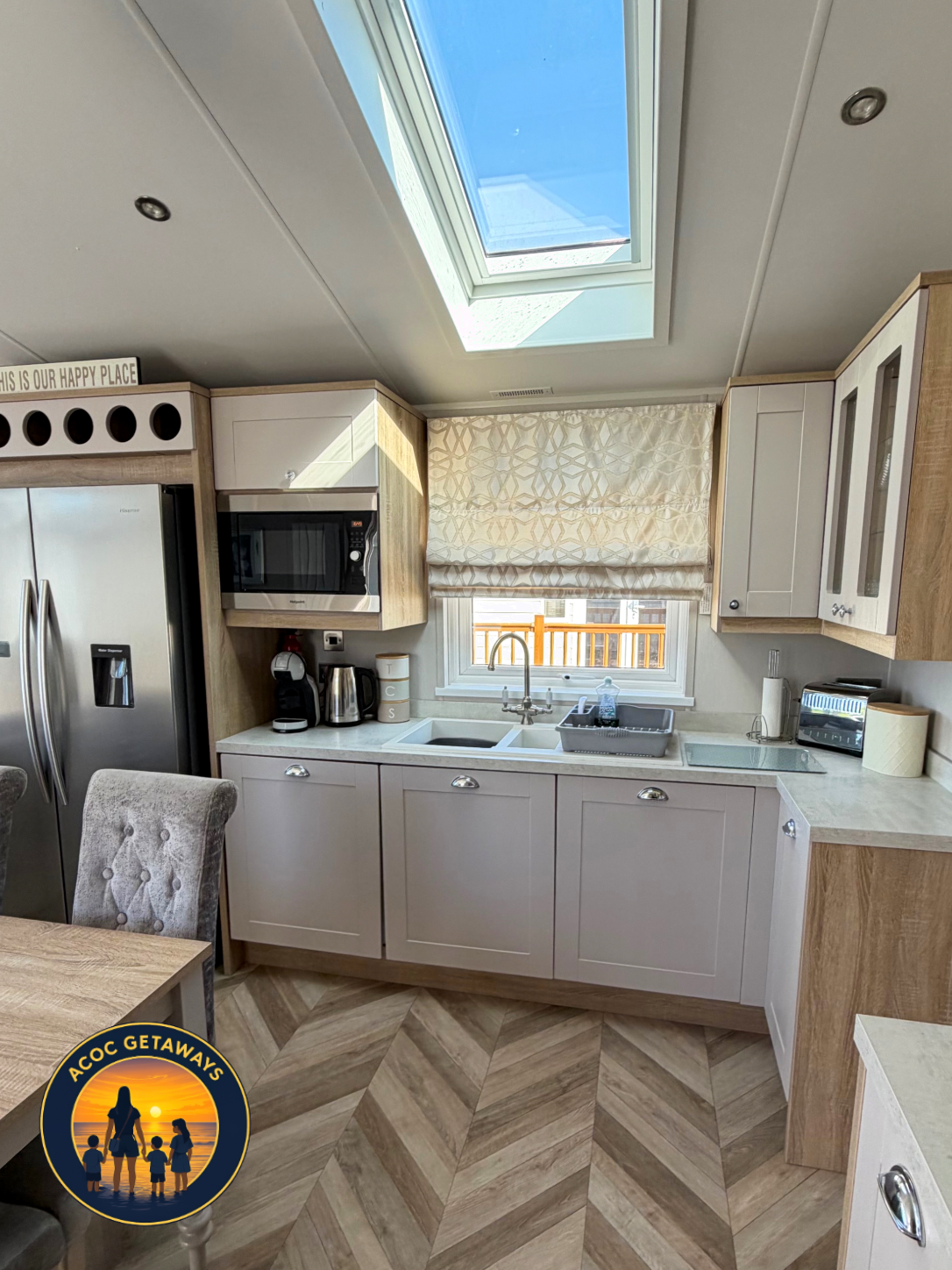 A modern kitchen with white and wooden cabinets, a skylight, a window with a patterned valance, a double sink, a coffee maker, microwave, toaster, and various kitchen items on the counters.