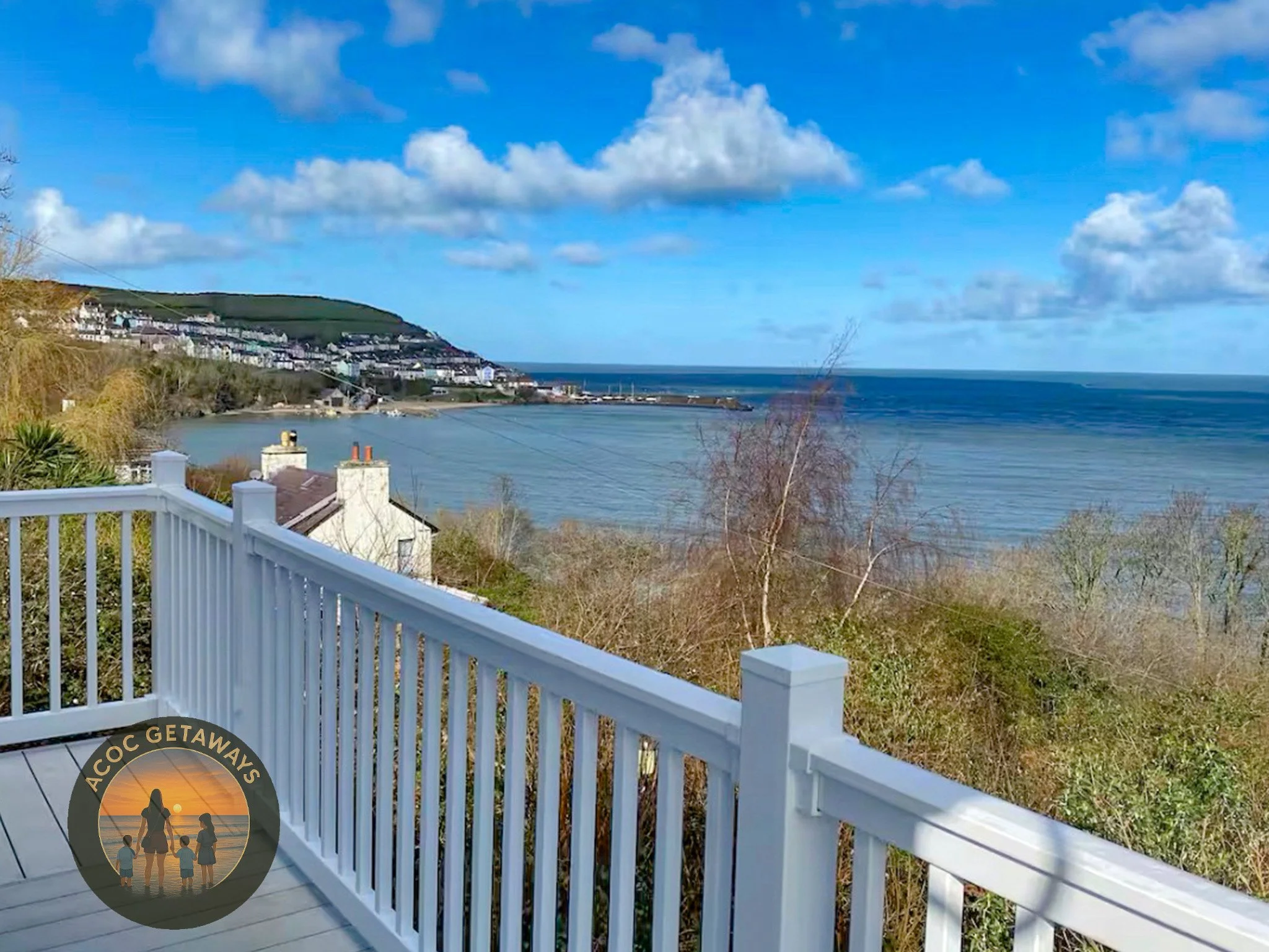 View from a balcony overlooking a coastal village with houses, a hill, and the ocean under a partly cloudy sky.