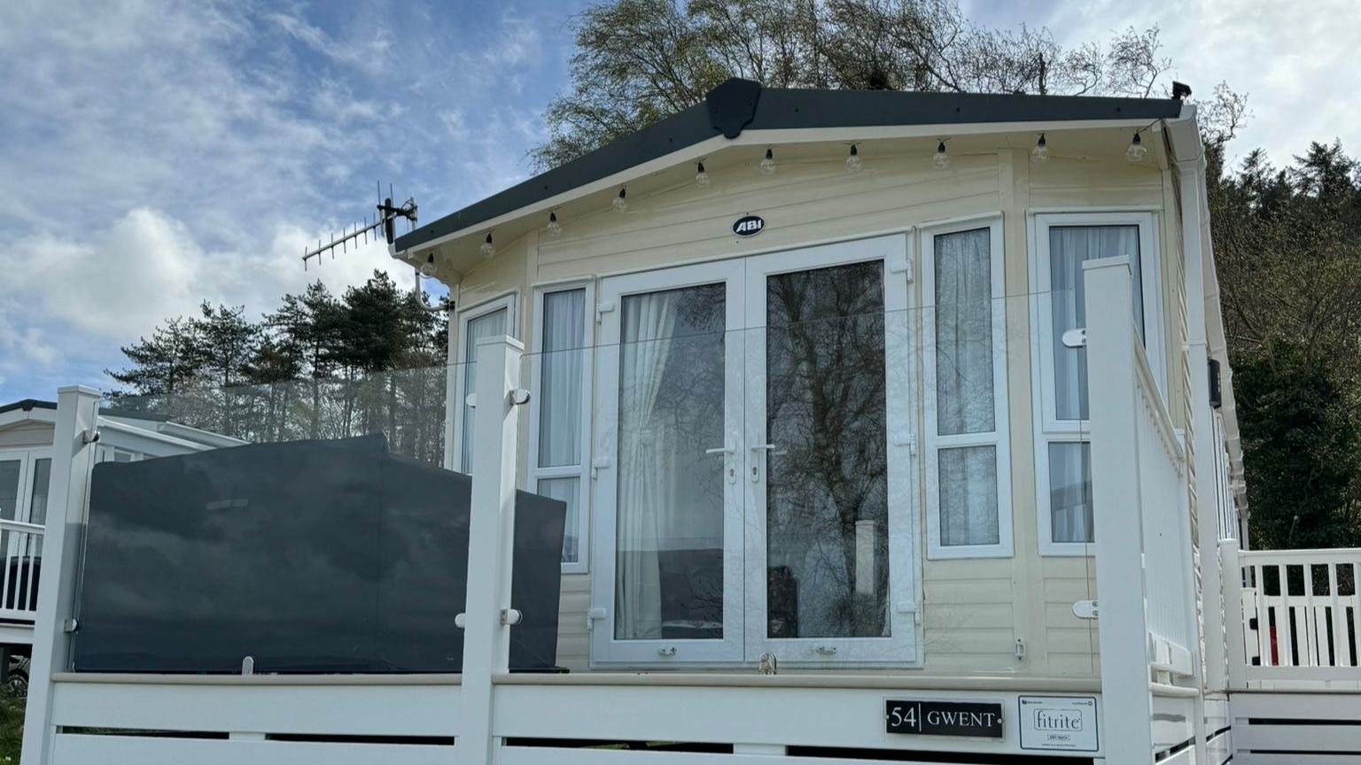 A small beige house with glass doors and multiple windows, surrounded by a white fence, with trees and a partly cloudy sky in the background.