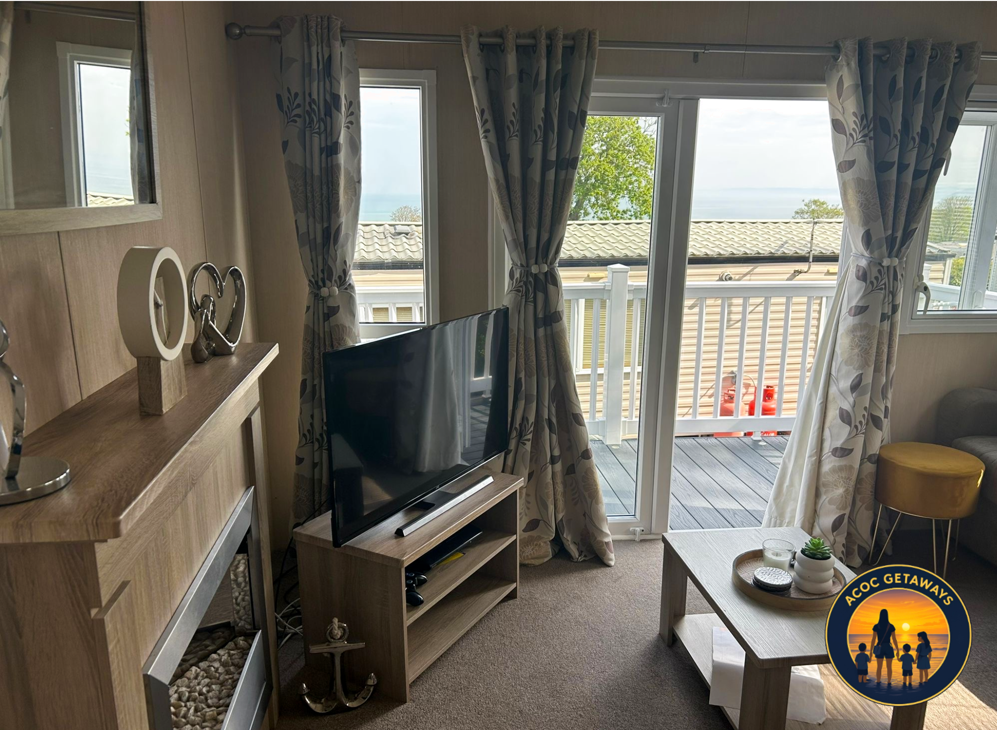Living room with large glass sliding door and window, patterned curtains, wooden furniture, a flat-screen TV, and a small table with decorative items, overlooking a balcony and an outdoor view with trees and rooftops.