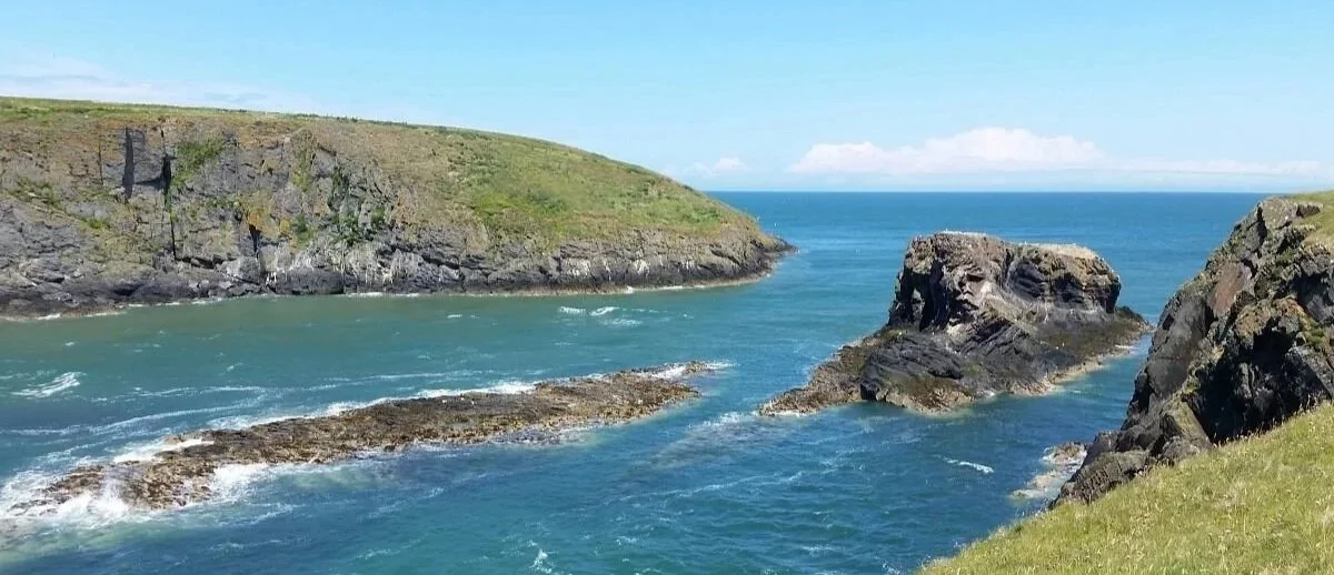 A rocky coastal landscape with cliffs, water, and grassy areas under a partly cloudy sky.