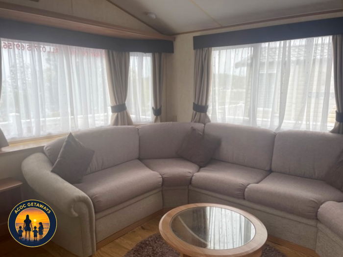 Living room with large beige sectional sofa, dark throw pillows, wooden floor, round glass coffee table, and large windows with curtains allowing natural light.