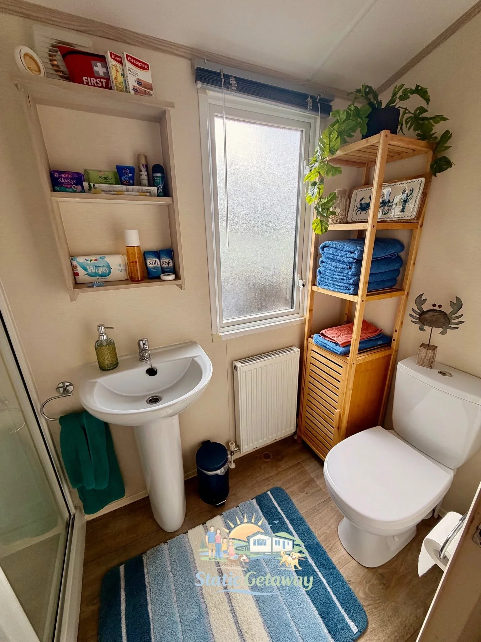 A small bathroom with a pedestal sink, a white toilet, a wooden shelf with folded blue towels, a potted plant on top, a frosted window with a blue blind, a small trash can, a wall-mounted rug with a beach scene, and a decorative crab on the wall.