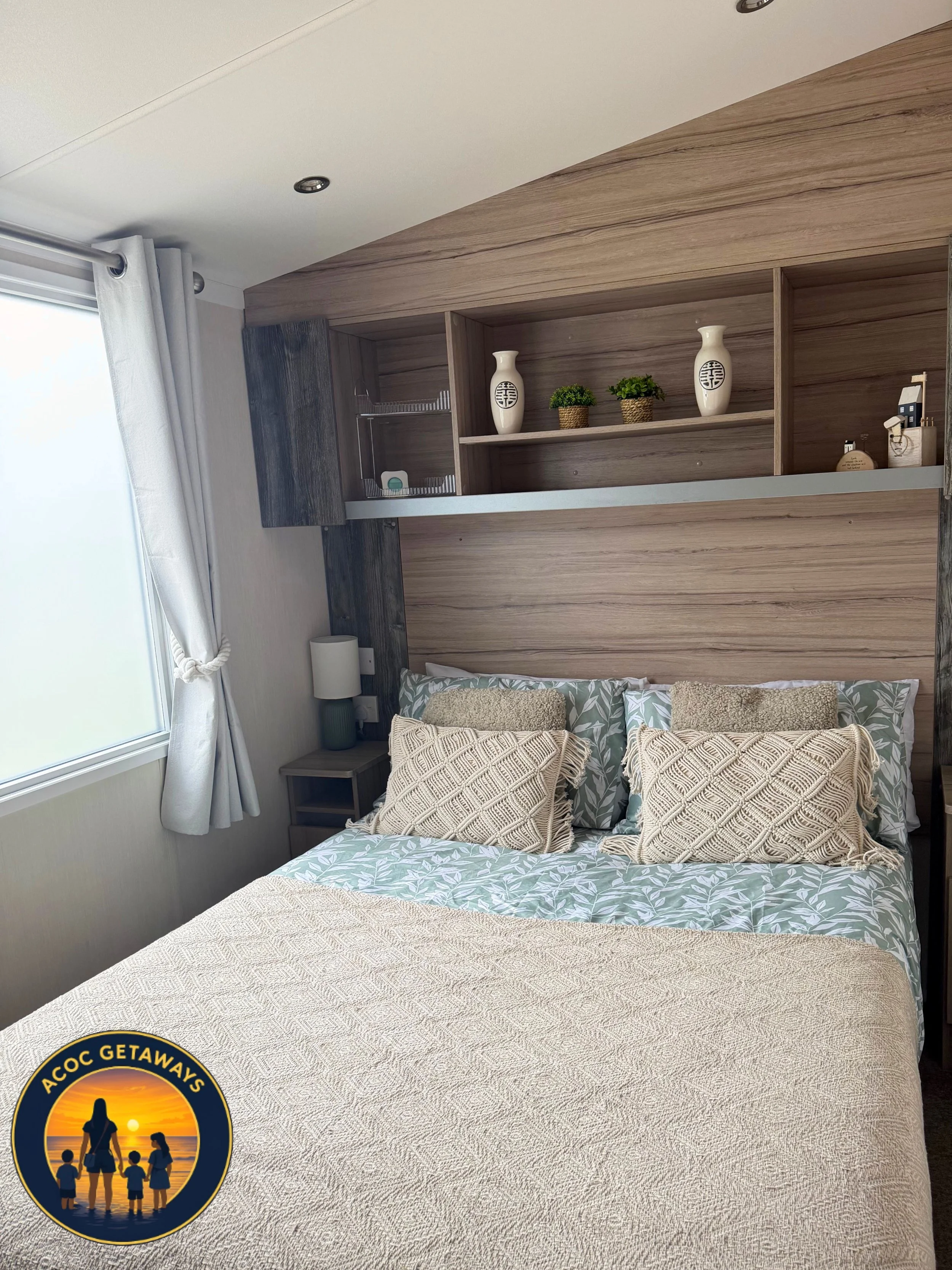 A bedroom with a large bed covered in a beige textured bedspread, decorated with patterned pillows. Behind the bed is a headboard with wood paneling and built-in shelves holding decorative vases and small plants. A window on the left with white curta