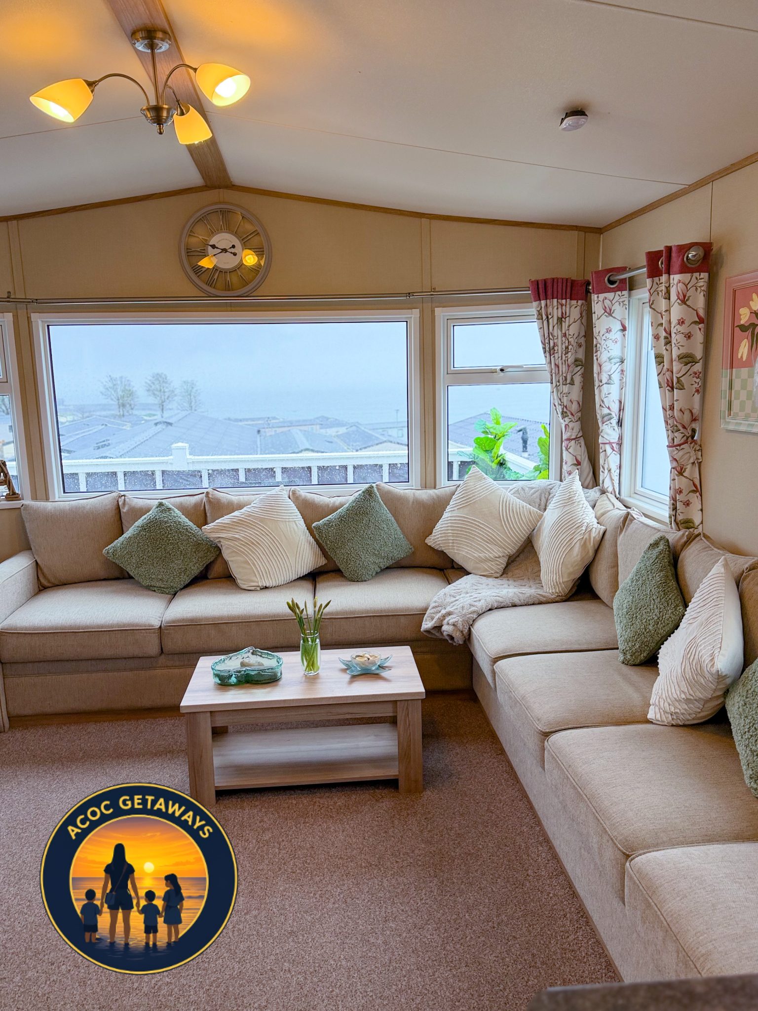 Living room with beige sectional sofa, multiple pillows, wooden coffee table, large window overlooking foggy landscape, floral curtains, ceiling light fixture, and wall clock.