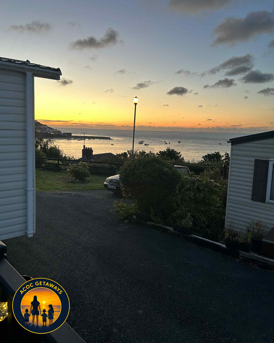 Sunset over a calm sea viewed from a residential area with houses and parked cars, streetlamp, and sky with scattered clouds.