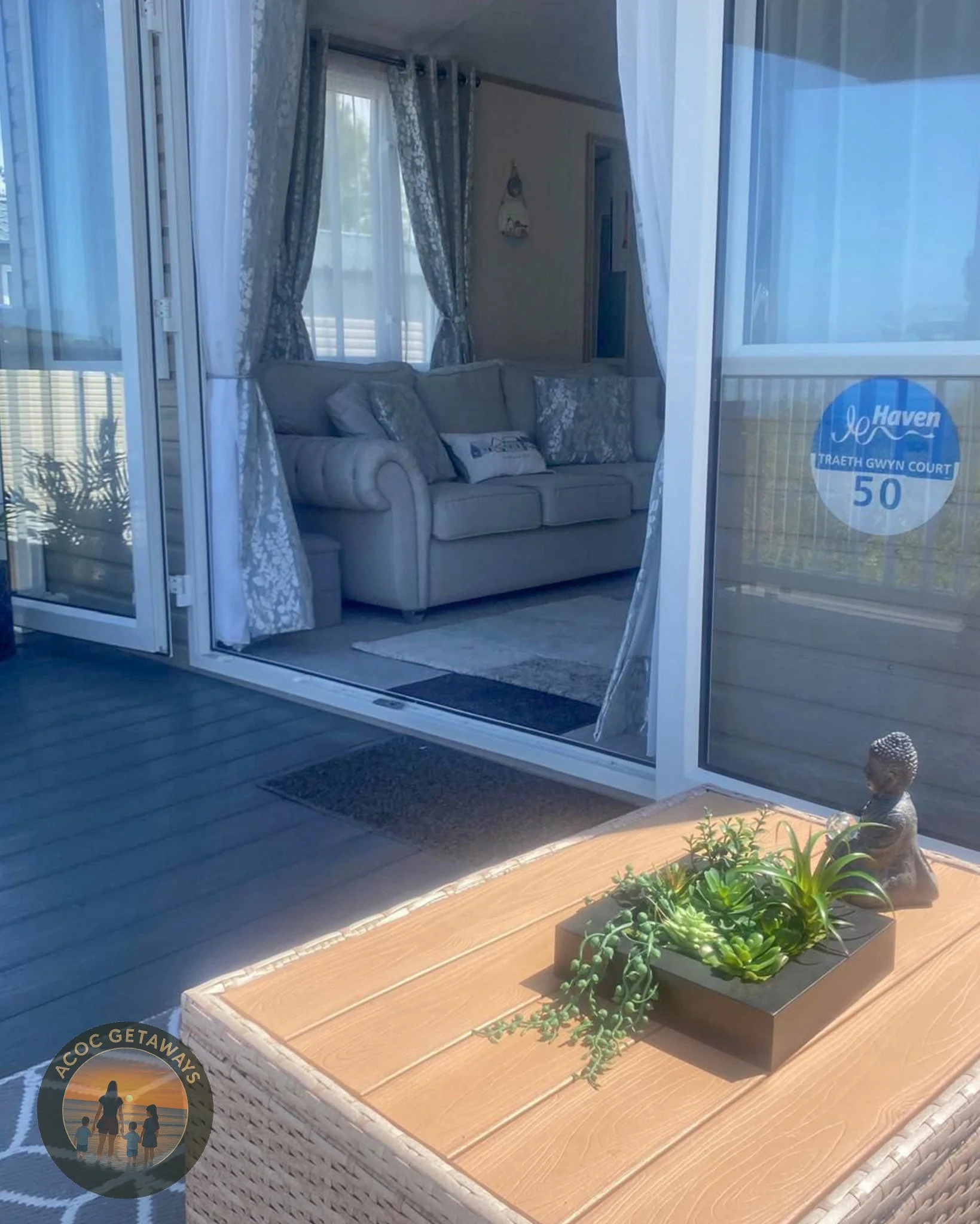 View through a glass door showing a living room with a beige sofa, curtains, and a wall hanging; a wooden table outside with a decorative plant and a Buddha sculpture; blue outdoor sticker on the door indicating address.