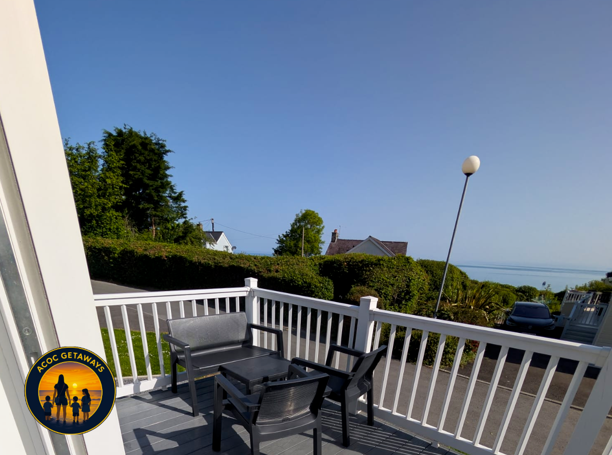 Outdoor patio with a table and four black chairs, white railing, hedge, trees, houses, streetlamp, parked car, and a view of the water in the distance on a clear day.