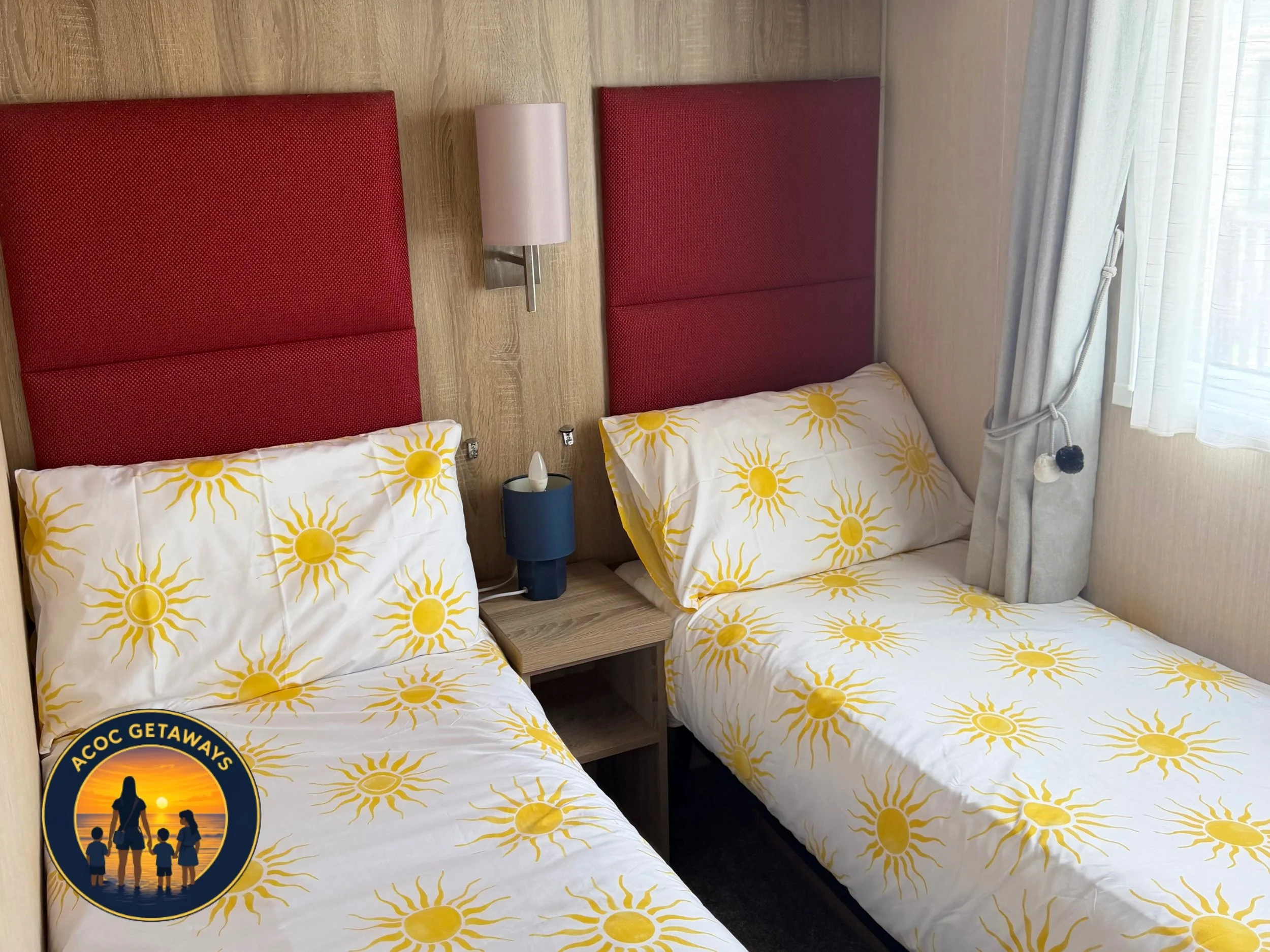 Two beds with yellow sunflower patterned bedding, red padded headboards, a blue nightstand with a white tissue box, a wall-mounted lamp, and a large window with translucent curtains.