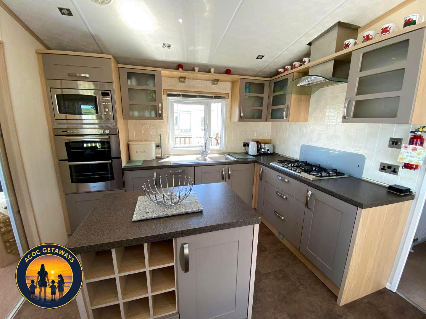 Modern kitchen with grey cabinets, black countertops, and appliances including a microwave, oven, and gas stove. Open shelving with red and white cups, and a small island with wine storage.