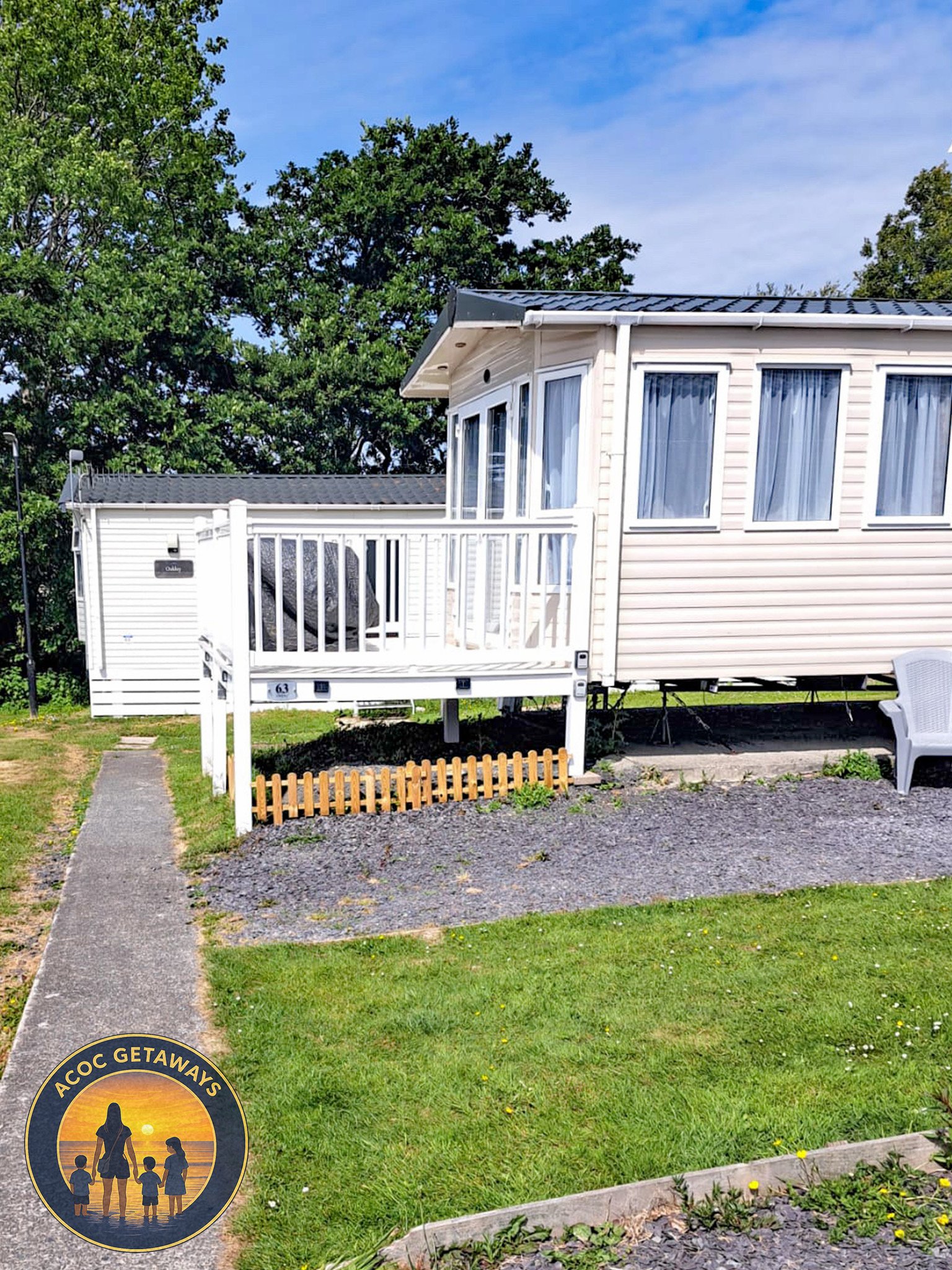 A beige mobile home with large windows, a small front porch with white railing, and a brown wooden fence in front. There is a gray chair on the right side and a pathway leading to the porch. Green grass and trees are visible in the background, and th