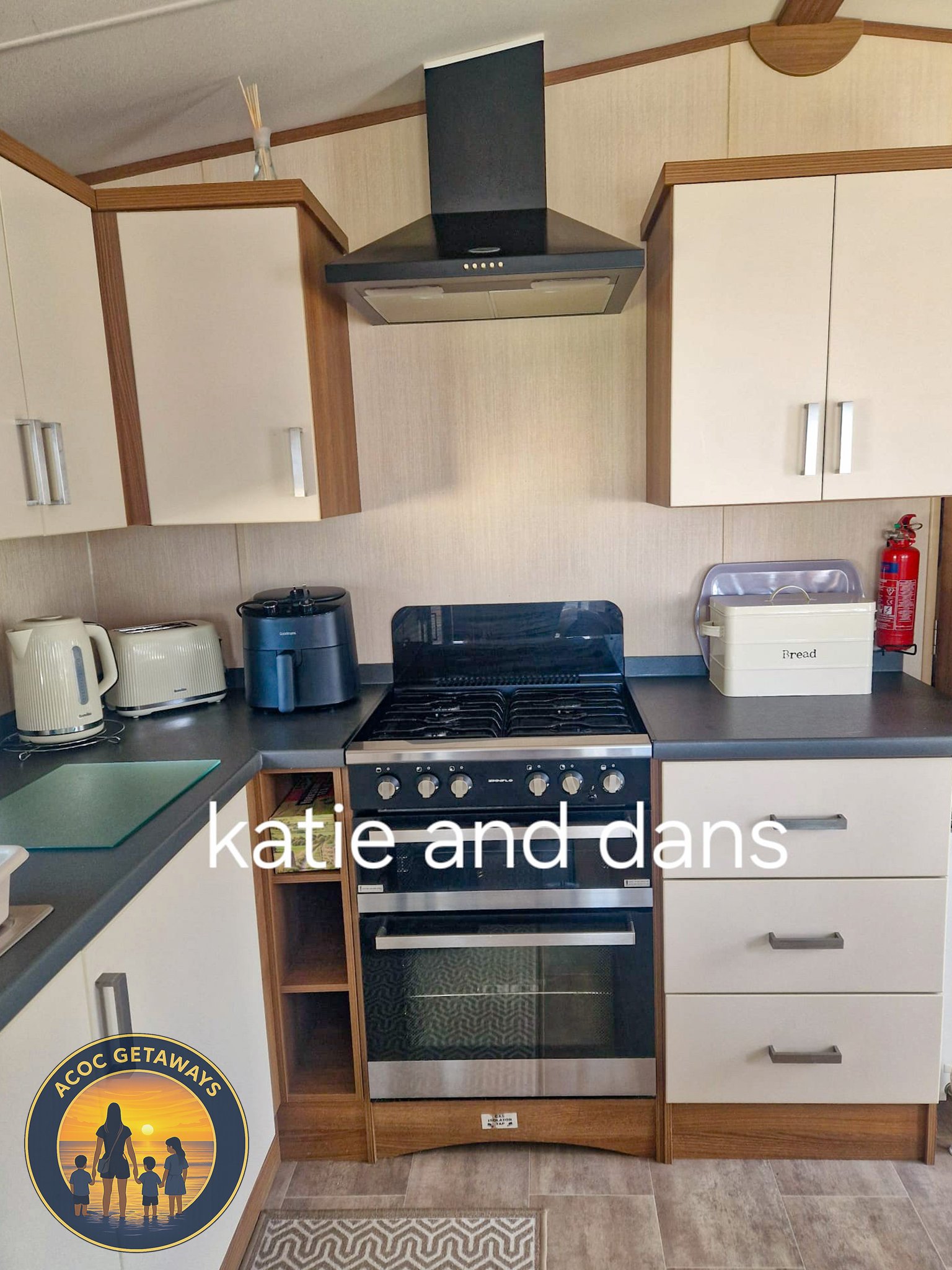 Kitchen with beige cabinets, a black stove with oven, a range hood, and various appliances on the counter including a kettle, toaster, and air fryer, with a fire extinguisher on the wall and a bread box.