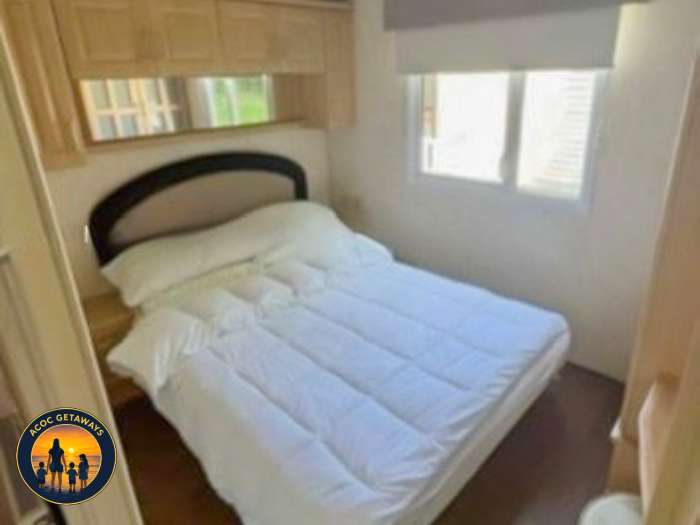 A neatly made bed with white bedding and pillows inside a small, bright room with a window.