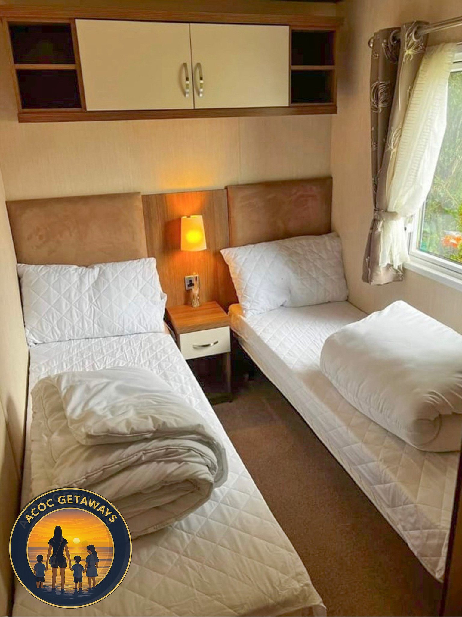 Hotel room with two single beds, pillows, rolled-up comforters, nightstand with a lamp, window with curtains, wall-mounted cabinet, and warm lighting.