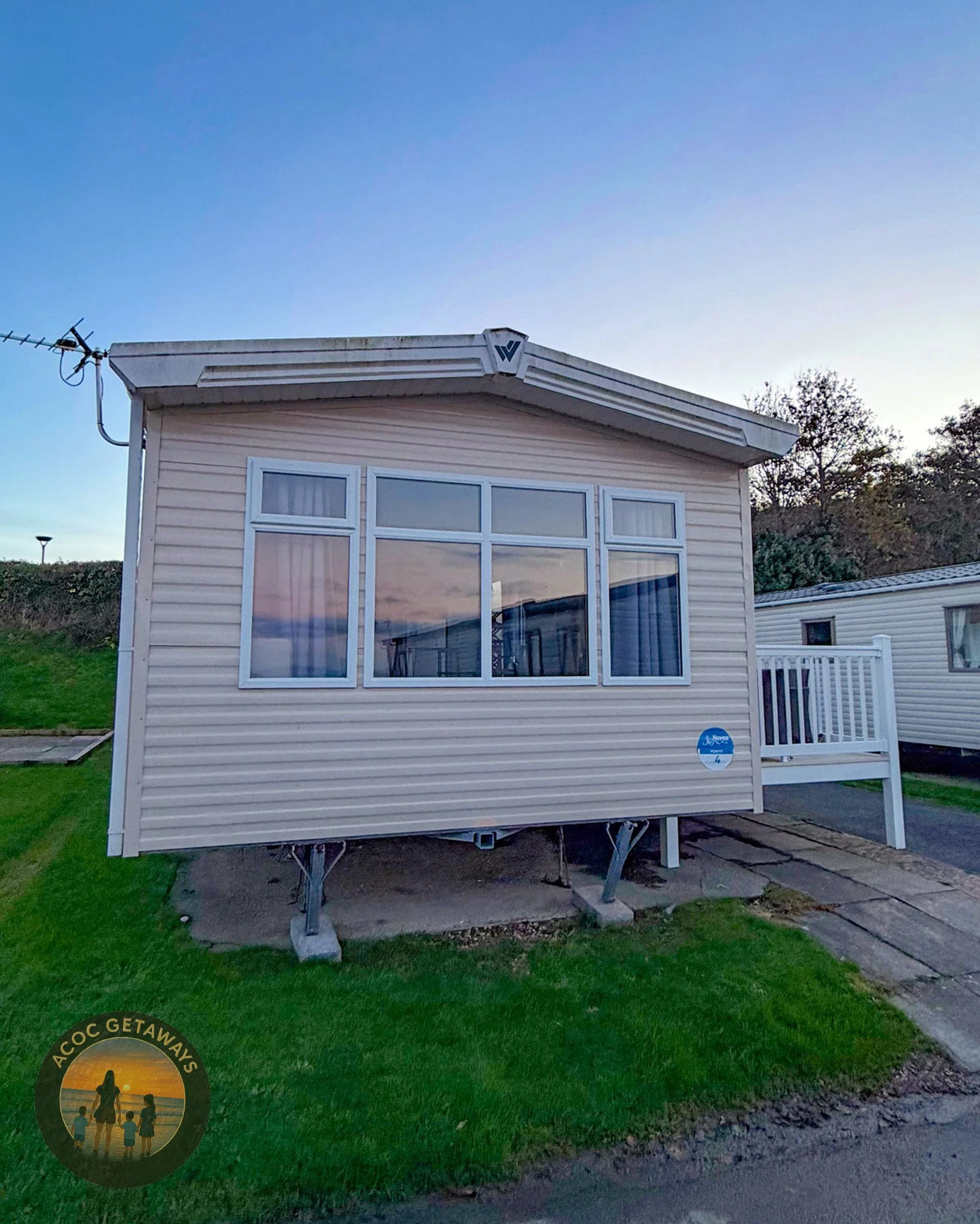 A small white mobile home with a raised entrance porch and a set of stairs, a large window reflecting the sunset, and green grass nearby, during dusk.