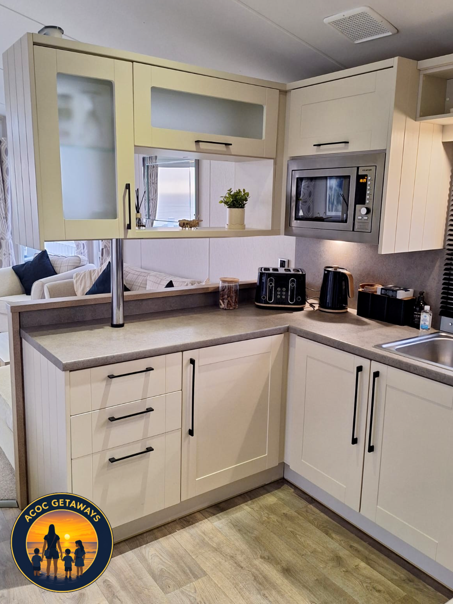 Kitchen with cream-colored cabinets, black handles, a microwave, a toaster, a kettle, and a small bottle of hand sanitizer on the counter. A mirror above the counter shows part of a living room with a sofa and cushions.