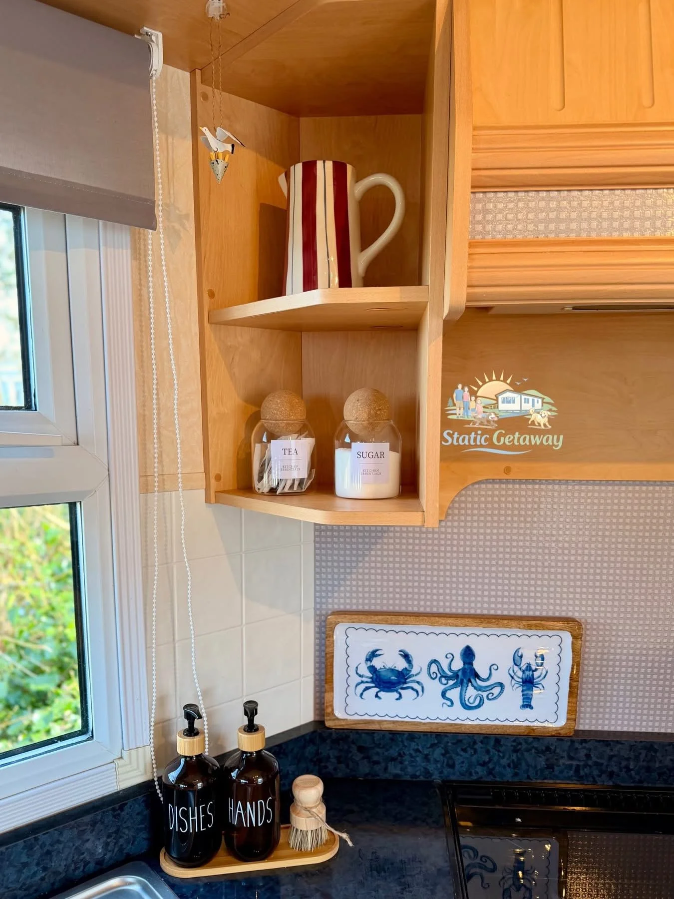 Kitchen bookshelf with a ceramic pitcher, two jars labeled 'Tea' and 'Sugar', and a decorative hanging bird. Below the shelf are bottles labeled 'Dishes' and 'Hands' with a dish brush and soap dispenser nearby. Part of a window and a framed crab artw