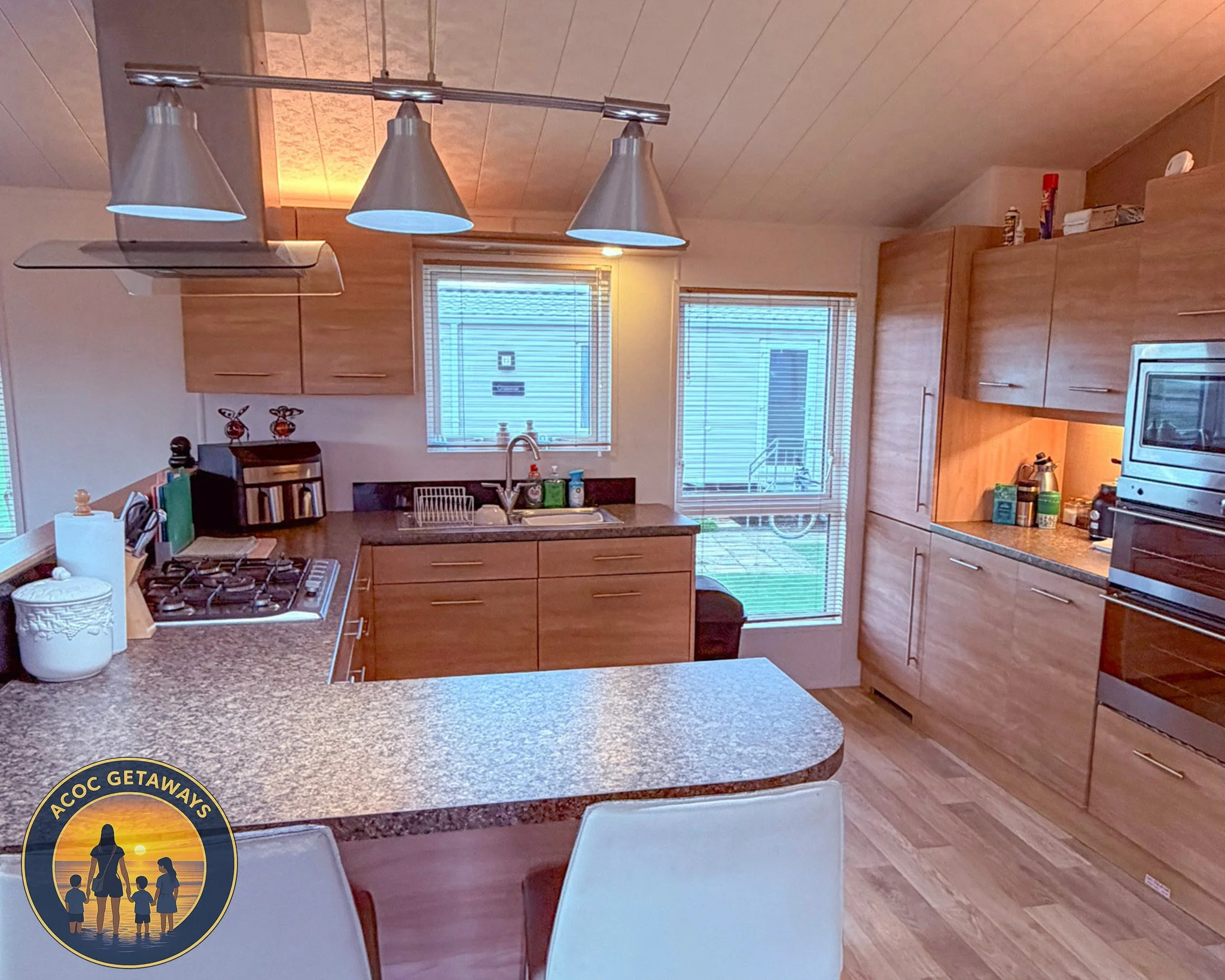 A kitchen with wooden cabinets, a granite countertop, a gas stove, and a stainless steel range hood. There are two windows with blinds, a microwave, various kitchen items, and a dining area with chairs. It has wood flooring and ceiling.
