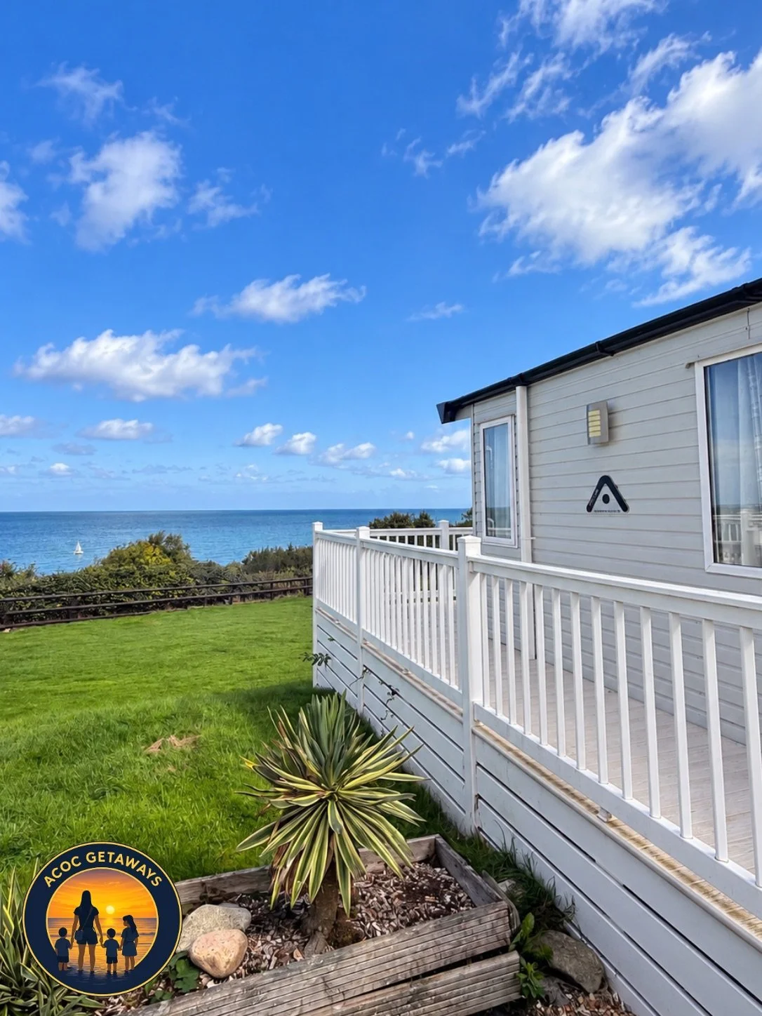 View of a beach house with a white deck railing, overlooking green lawn, shrubs, ocean, and a blue sky with scattered clouds.