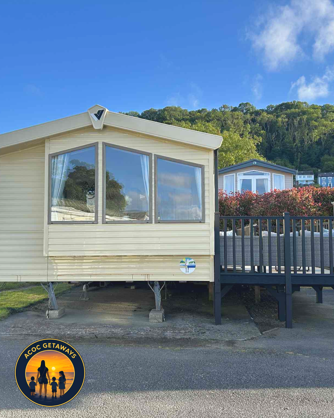 A beige mobile home with large front windows, a small deck with black railing, and a logo in the bottom left corner reading 'ACOC Getaways' with silhouettes of a family at sunset.