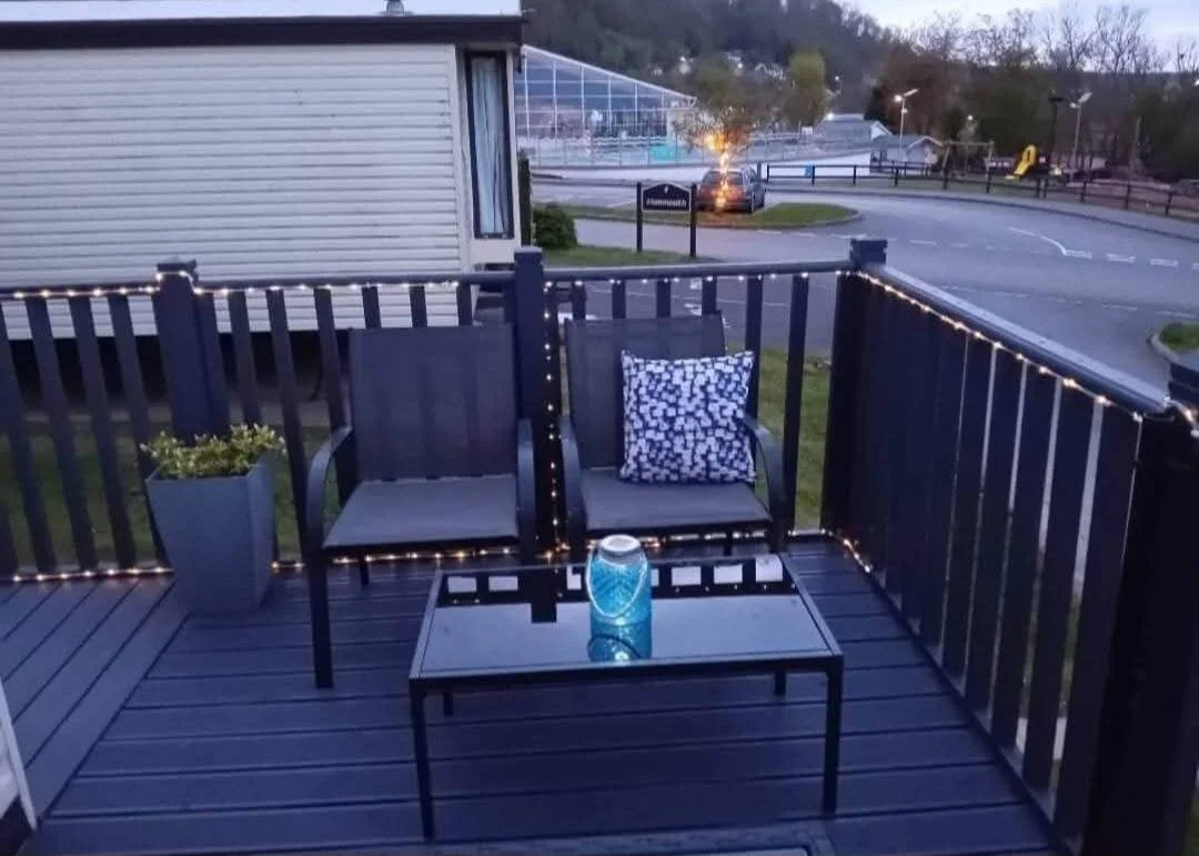 Small balcony with black wooden chairs, a table with a blue candle, a cushion, potted plant, and string lights, overlooking a parking lot and street.