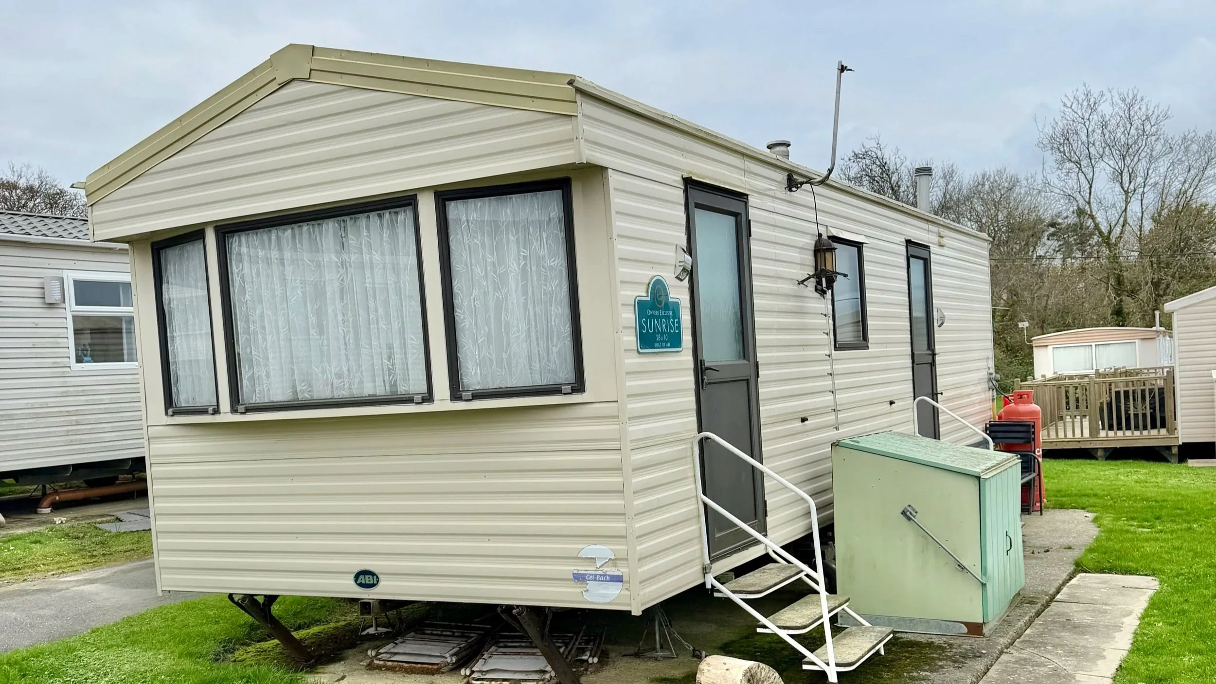 A beige mobile home with black window frames and steps leading up to the door, situated on a grassy area with trees in the background.
