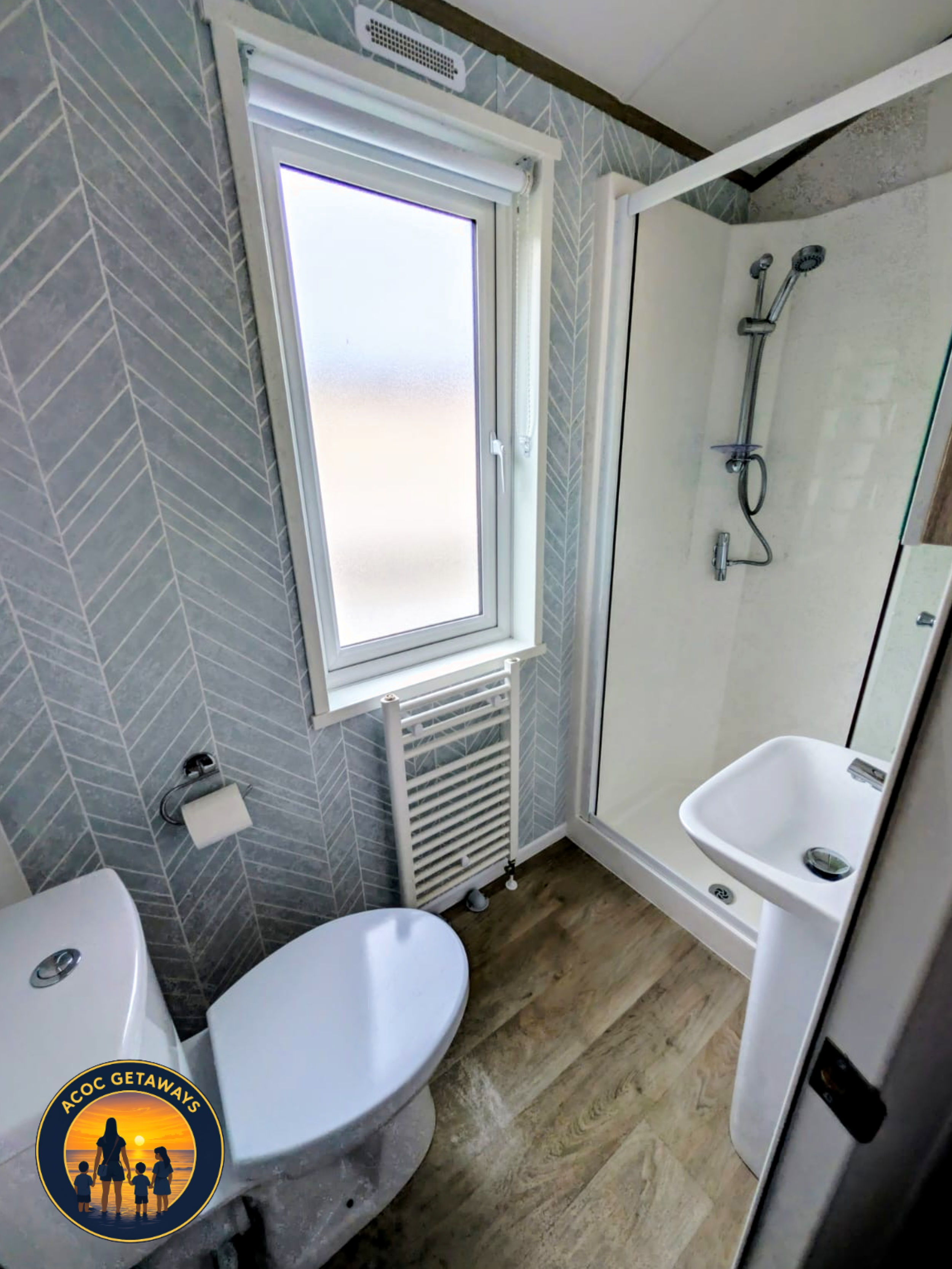 A small bathroom with a modern toilet, a sink, a shower with a glass door, a tall thin window, a radiator under the window, and a toilet paper holder. The floor is wood-look laminate and the walls are decorated with gray tiles arranged in a herringbo