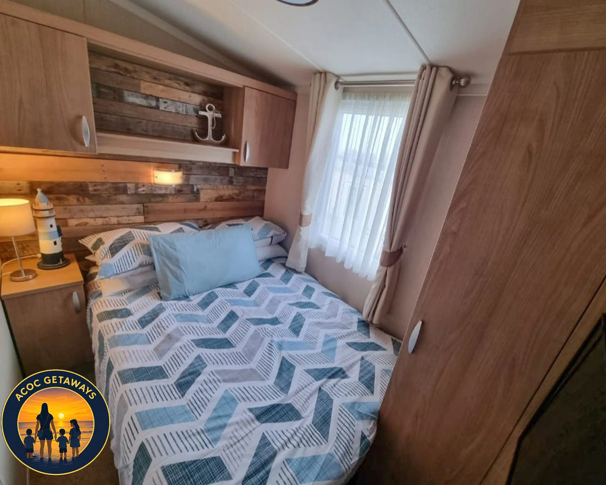 A cozy bedroom with a neatly made bed featuring a geometric patterned quilt in shades of blue, white, and gray, with a light blue pillow. There is a wooden headboard with a rustic wood plank design and a small living anchor decoration. To the left, a