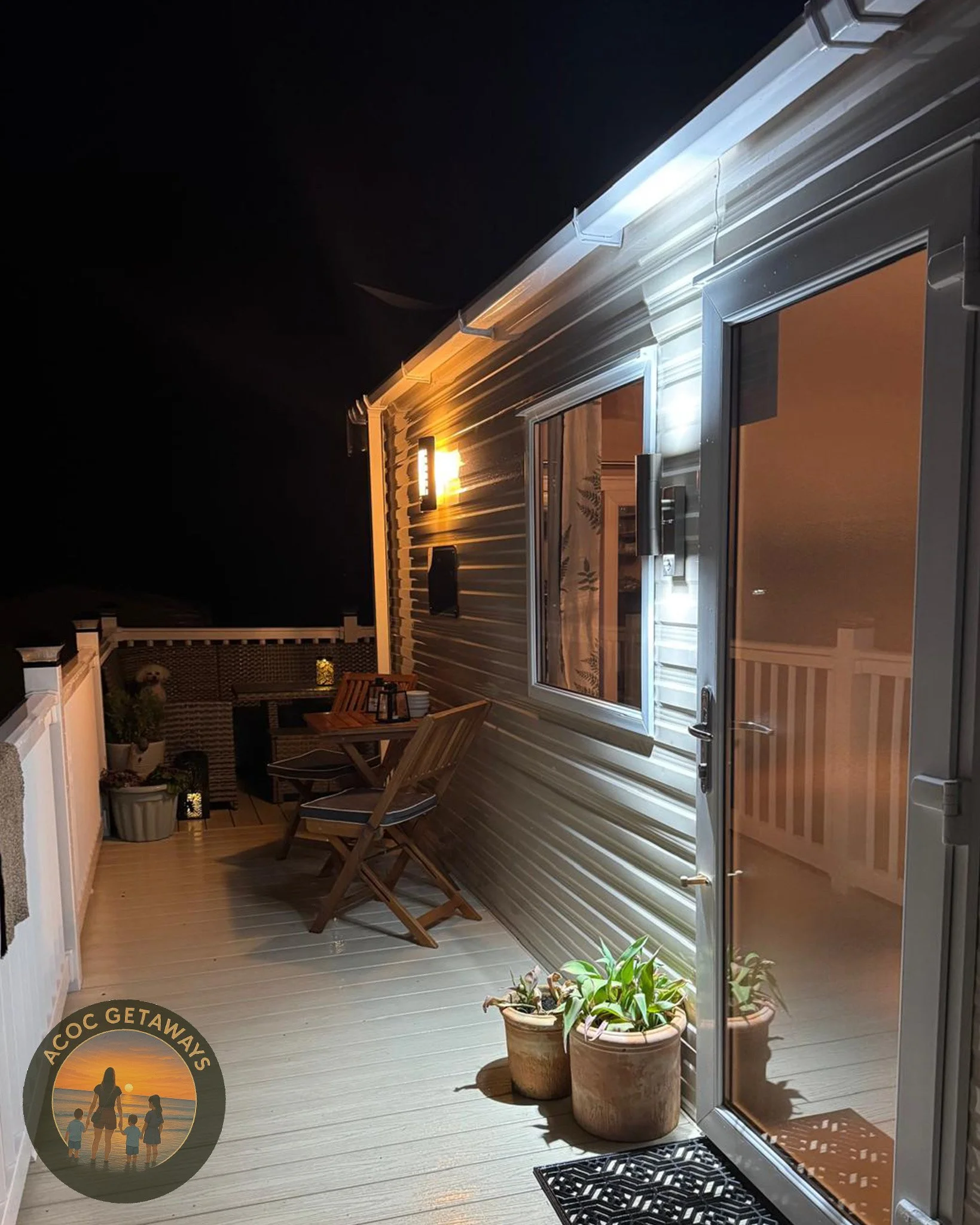 Nighttime view of a cozy outdoor balcony with wooden furniture, potted plants, and warm lighting, overlooking a dark sky.